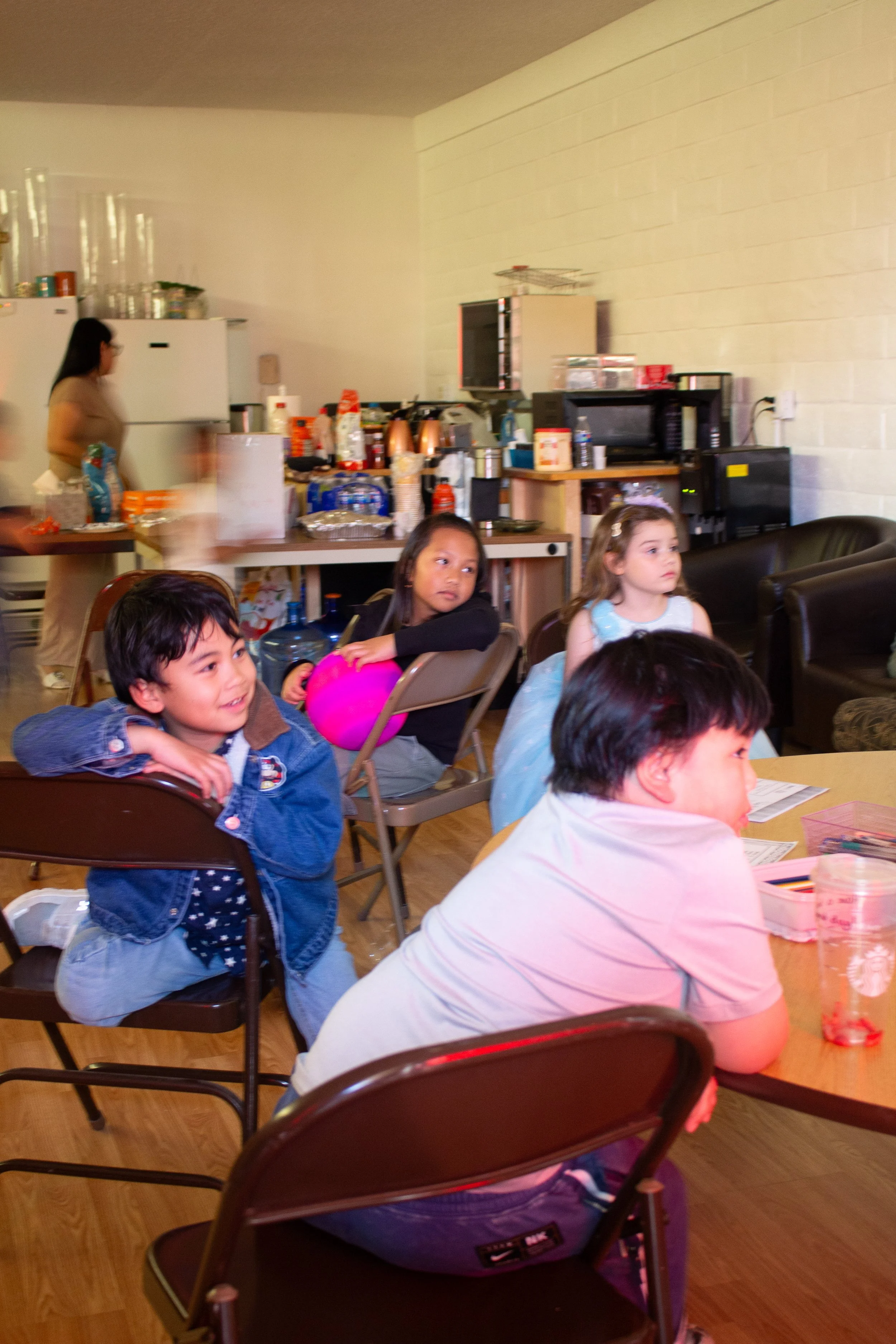 Children sitting at a table with snacks and drinks during a casual indoor gathering, with a woman in the background near a kitchen area.