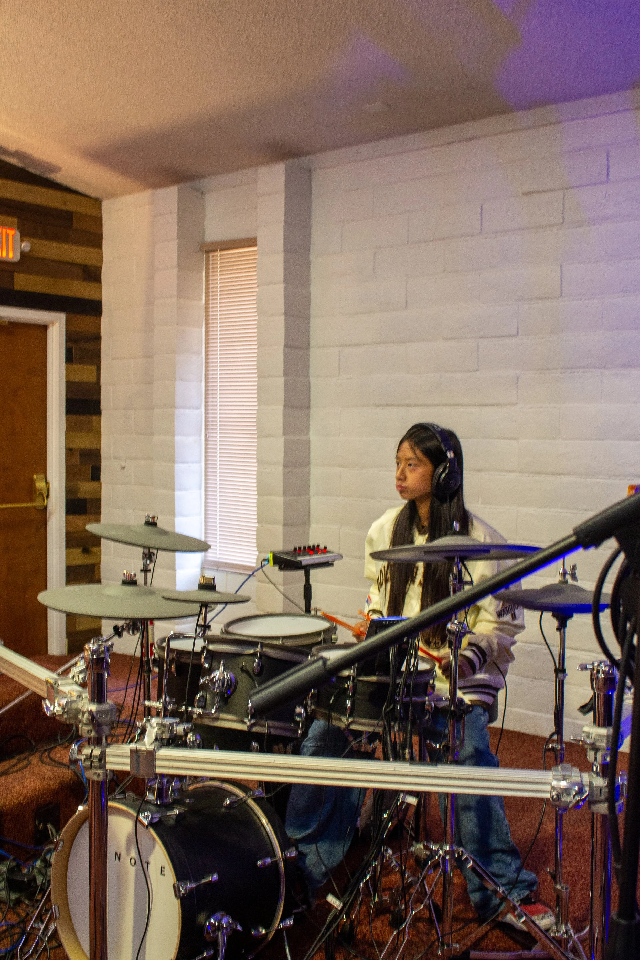A young girl wearing headphones is sitting behind a drum set on a stage in a room with white brick walls. There is a window with blinds and a wooden door with an exit sign.