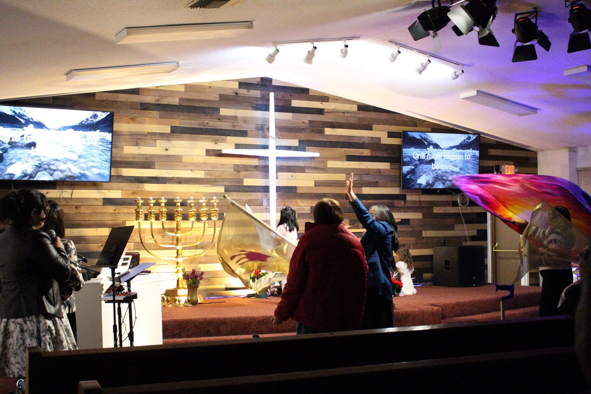 Inside a church or auditorium with people, a large cross and menorah at the front, two screens displaying a mountain landscape, a woman playing keyboard, and an individual raising their hand.