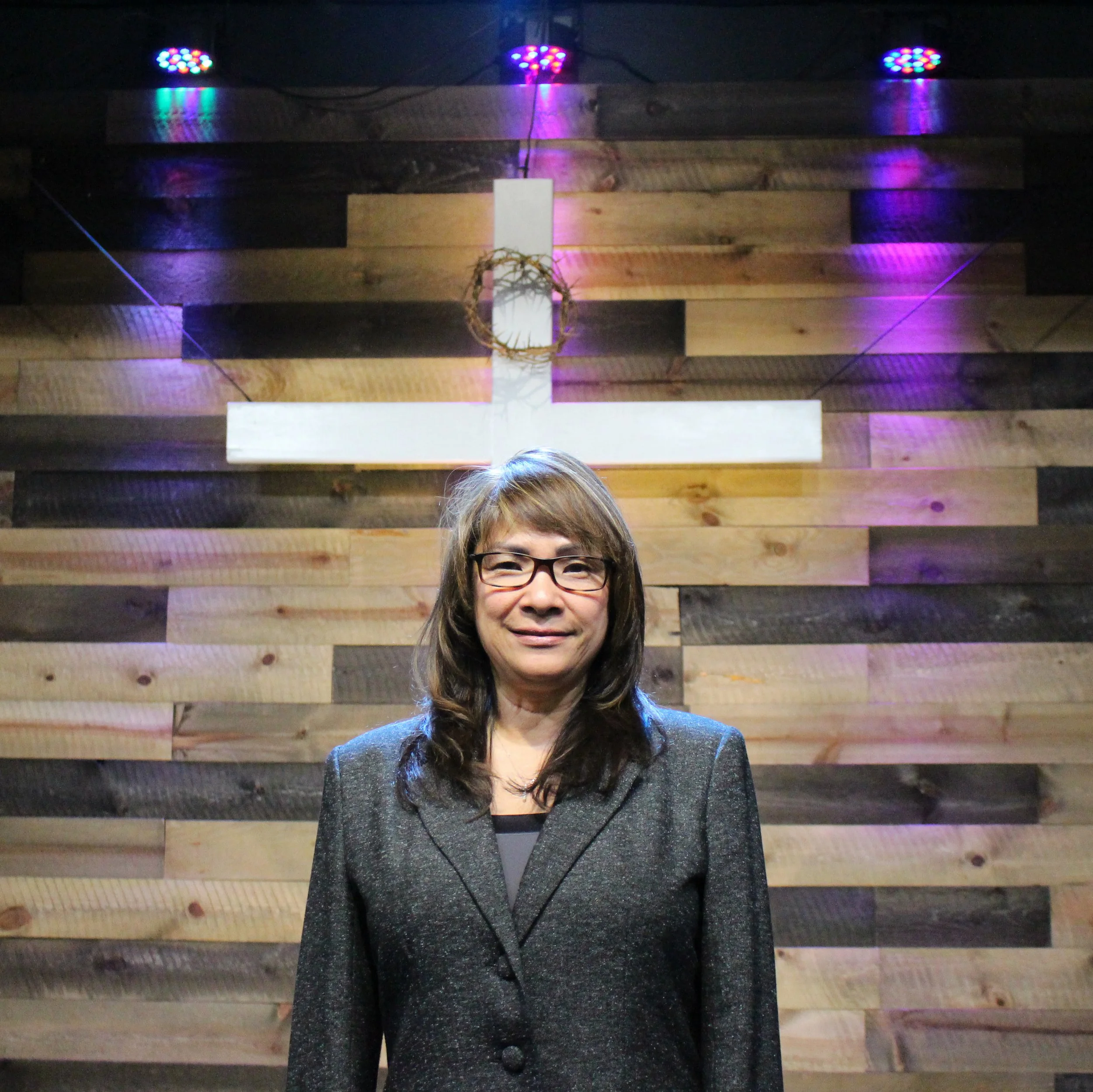 A woman with long dark hair and glasses stands in front of a wooden wall with purple and blue lights casting reflections on it.