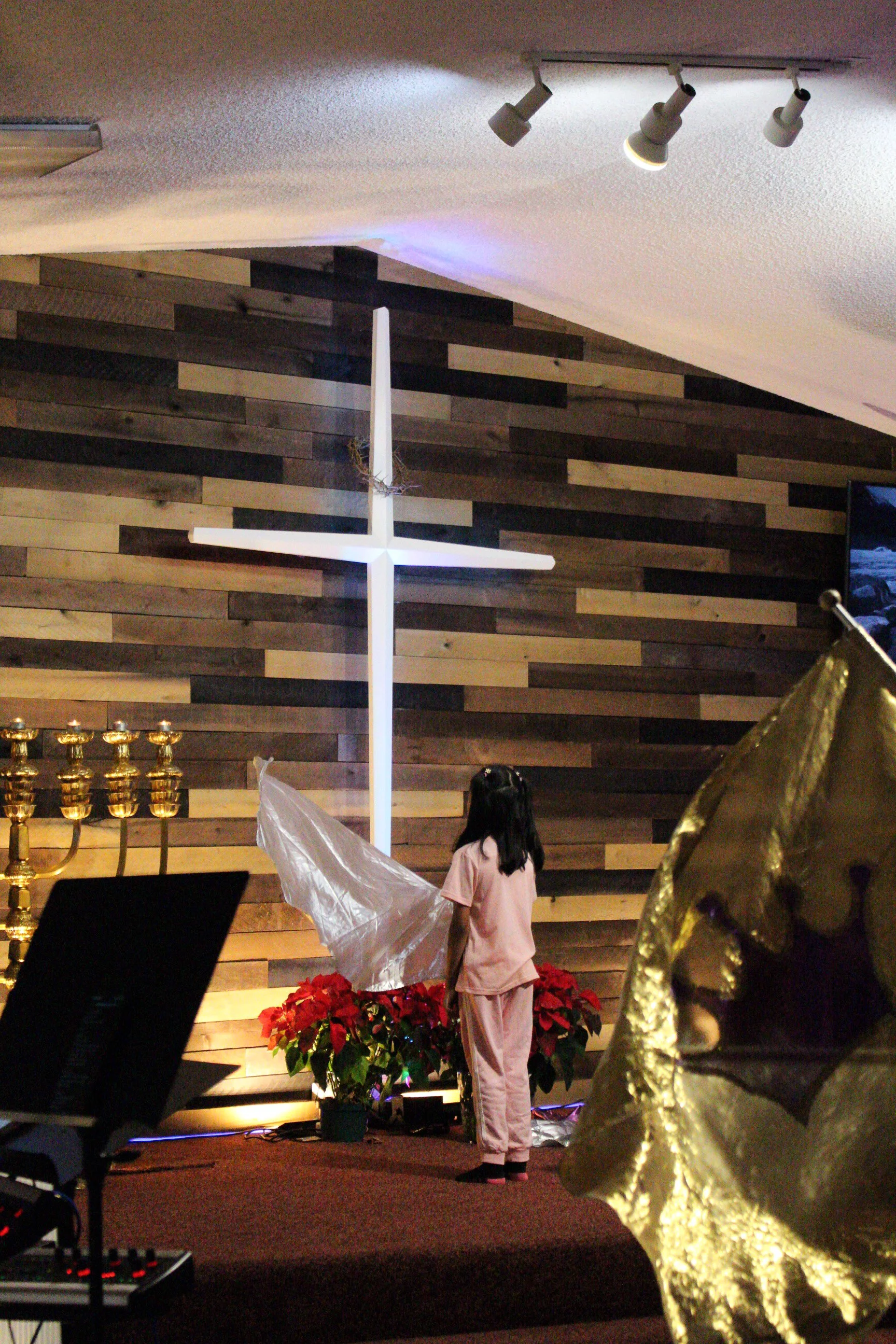 A person wearing pink pajamas standing next to poinsettia plants, inside a room decorated for Christmas with a wooden wall, a cross, Christmas lights, and a large gold angel statue.