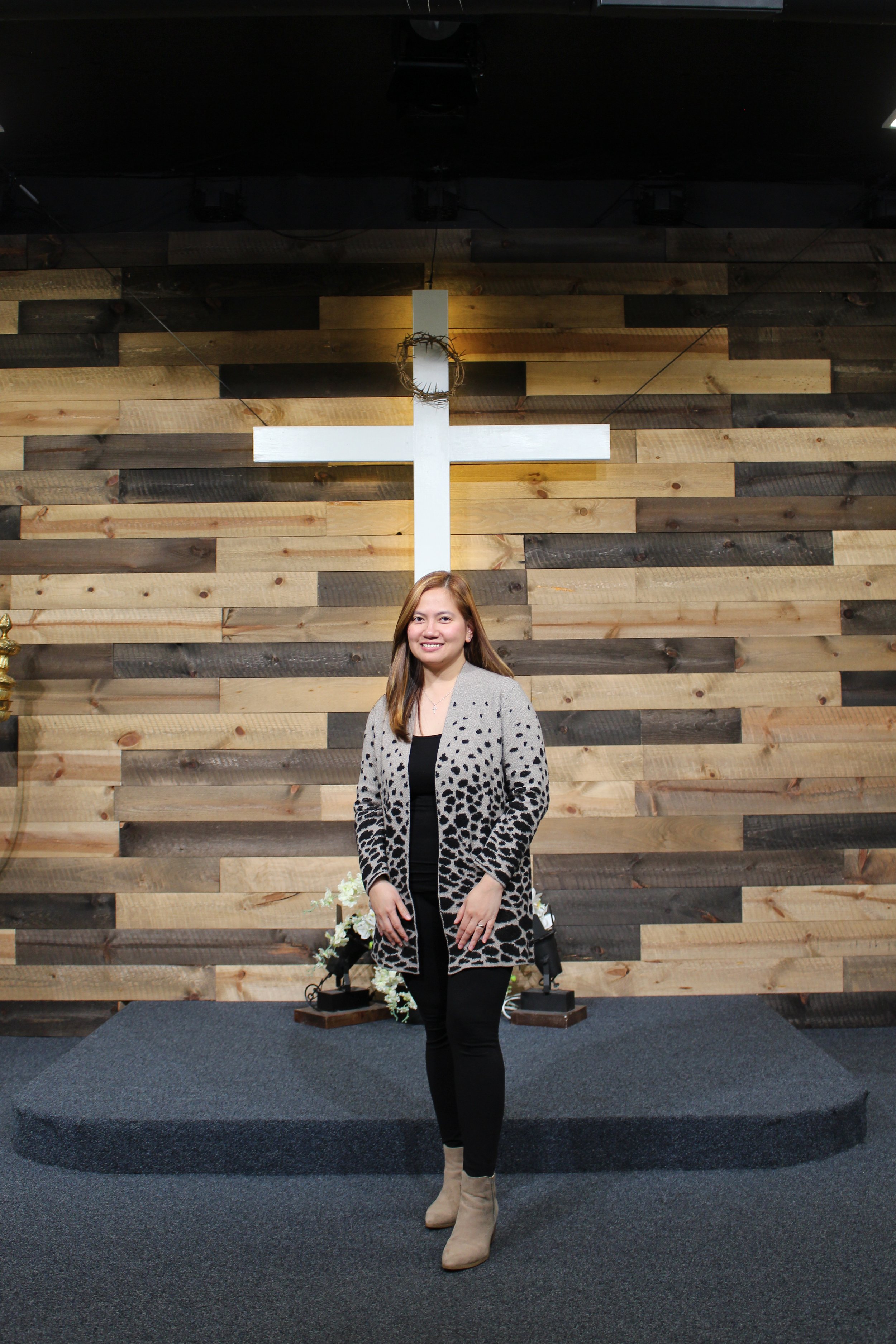 A woman standing on a stage in front of a wooden accent wall with a white cross and a crown of thorns above it. She is smiling, wearing a leopard print cardigan over a black top, black pants, and beige ankle boots.