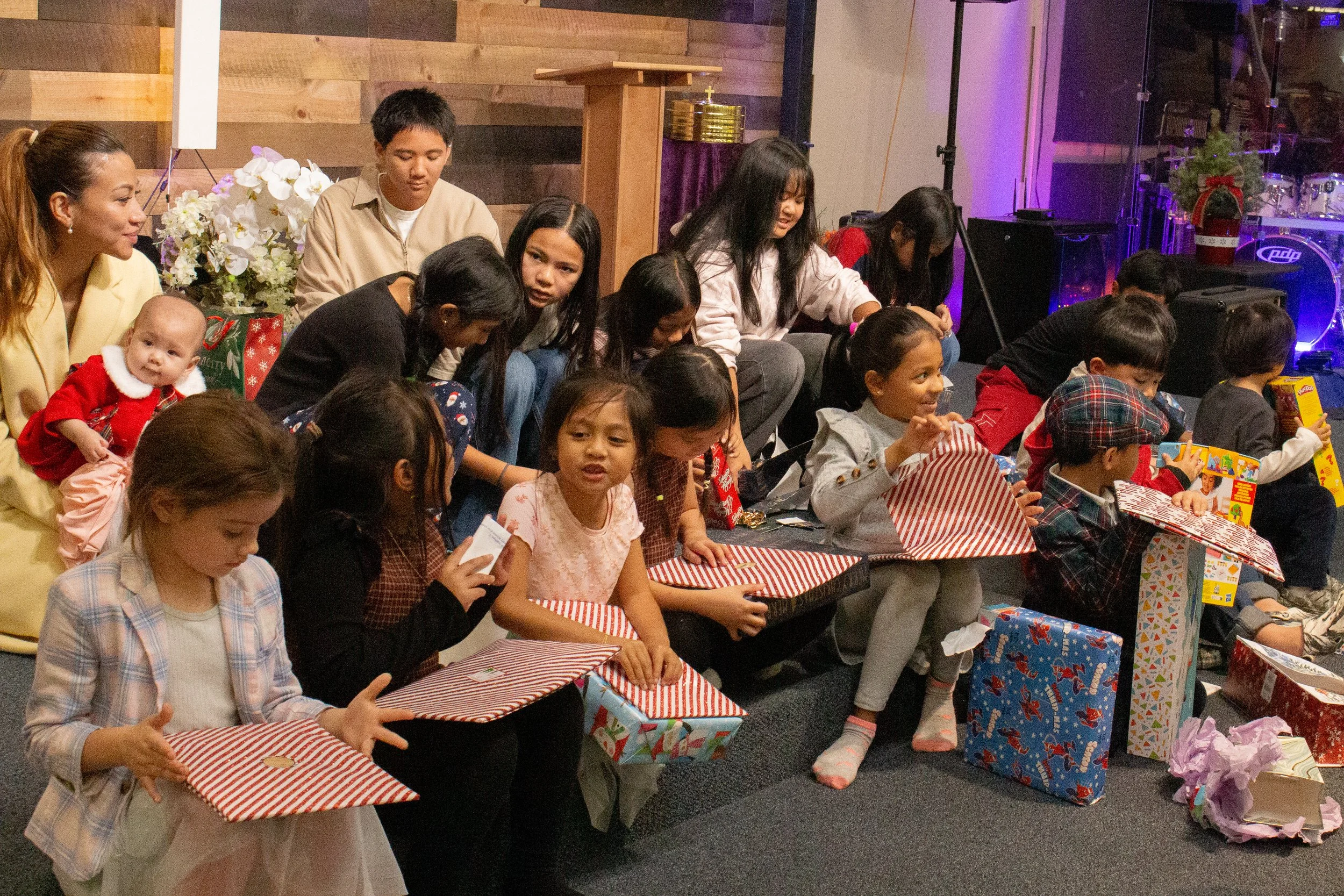 A group of children and adults opening Christmas presents on a stage, with Christmas-themed wrapping paper and gifts around them.