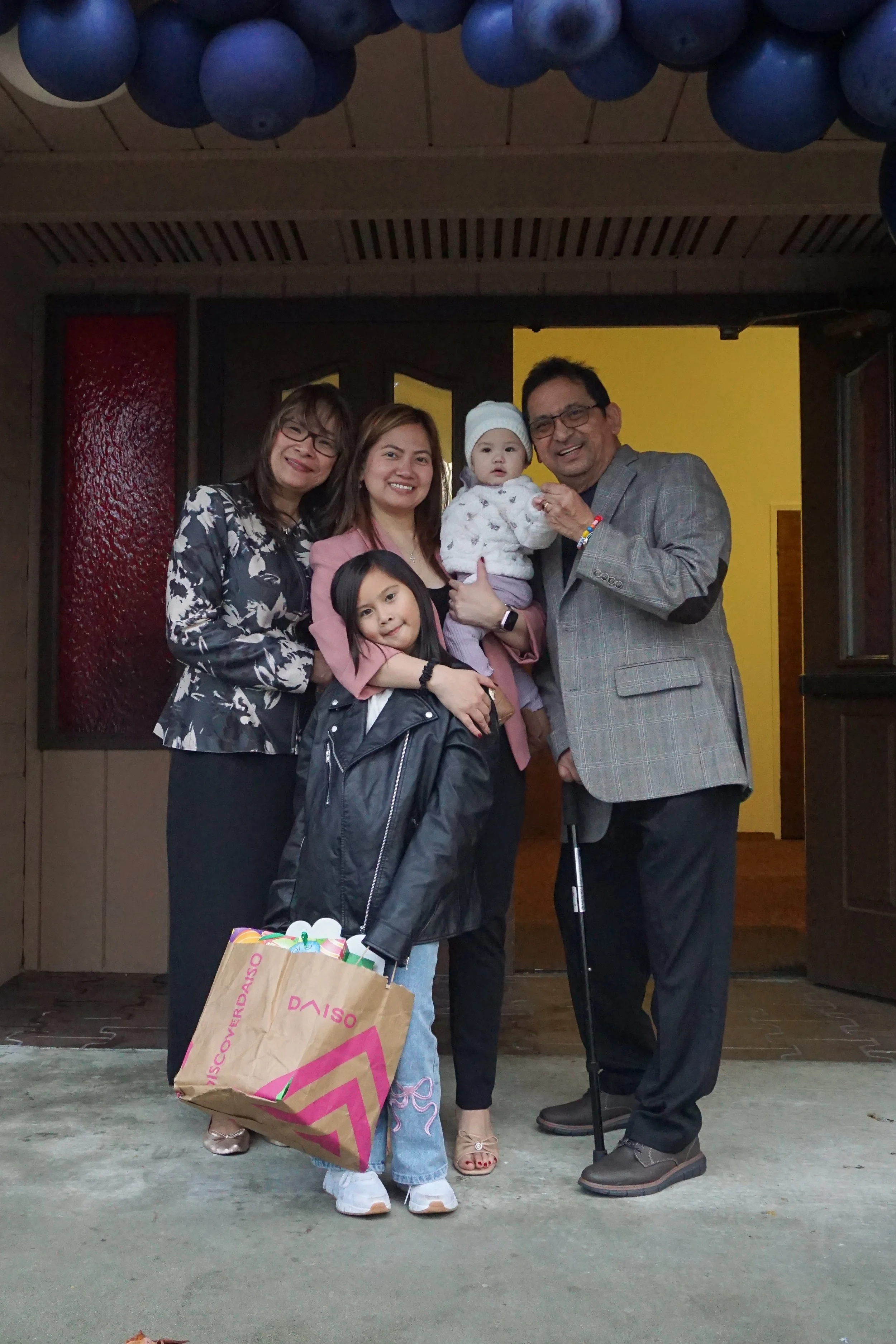 A group of five people, including two children and three adults, standing in front of a yellow door and a red window, smiling for the camera. One woman is holding a young child, and a girl in a black leather jacket is holding a shopping bag from Daiso.