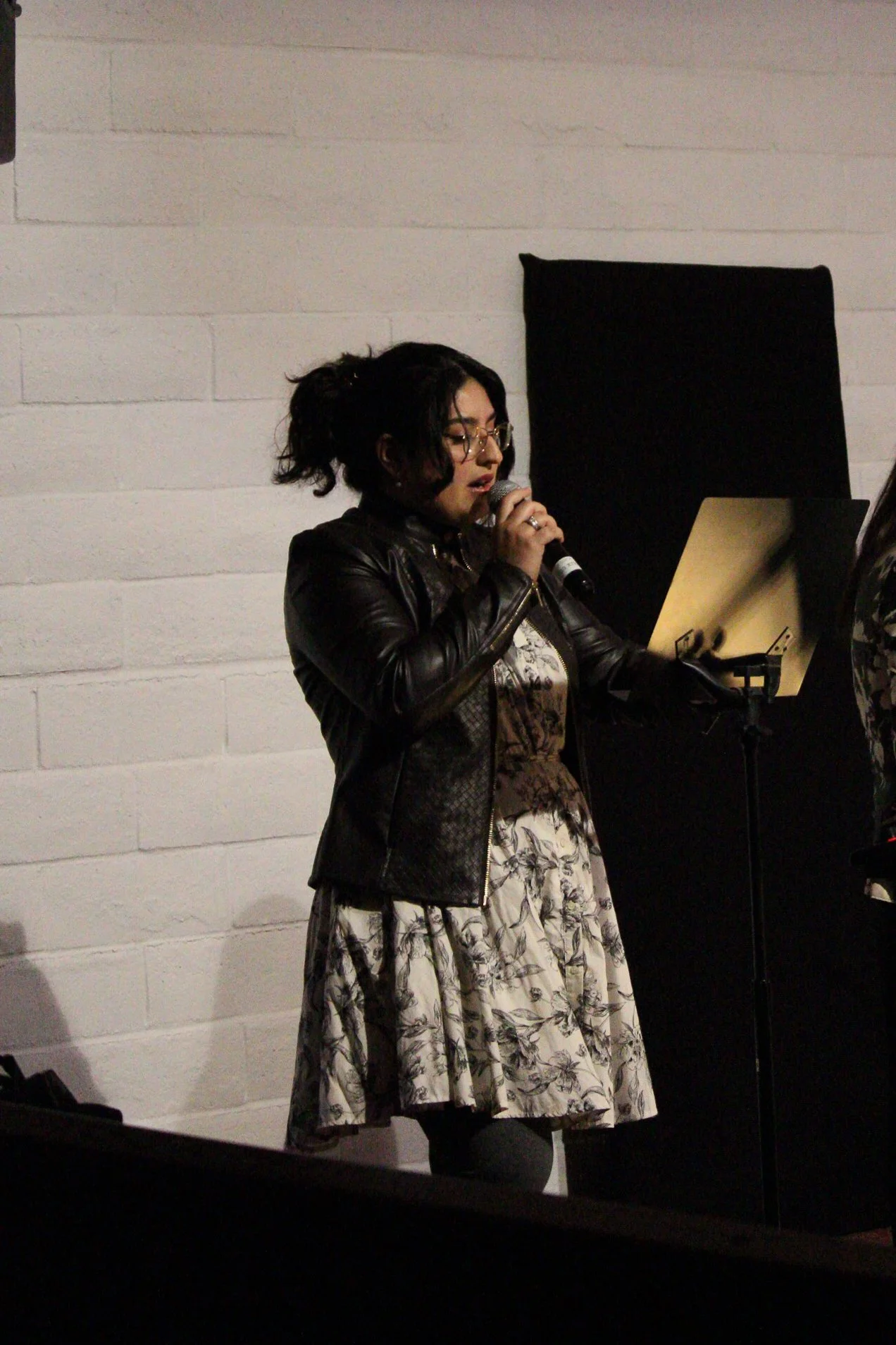 A woman with glasses and dark hair styled in loose curls is singing into a microphone while reading sheet music from a music stand. She is wearing a black leather jacket over a floral dress. The background features a white brick wall.