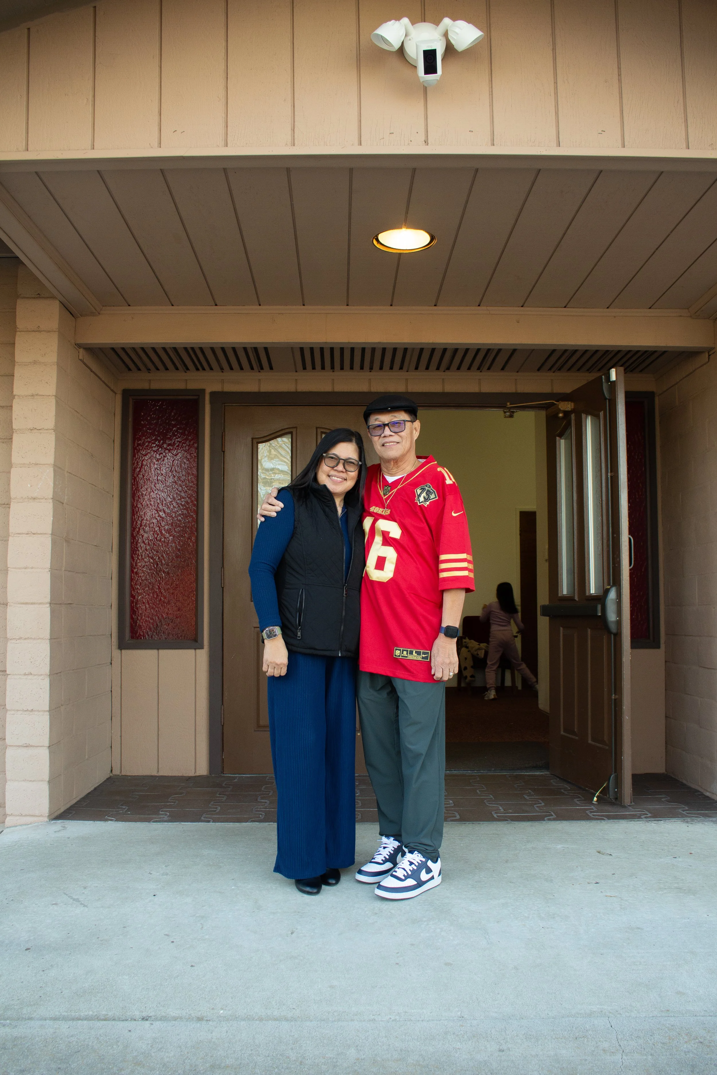 A woman and a man are standing together outside a house, smiling at the camera. The woman is wearing glasses, a black vest over a blue outfit, and black shoes. The man is wearing glasses, a black cap, a red sports jersey with the number 16, gray pants, and white and black sneakers.