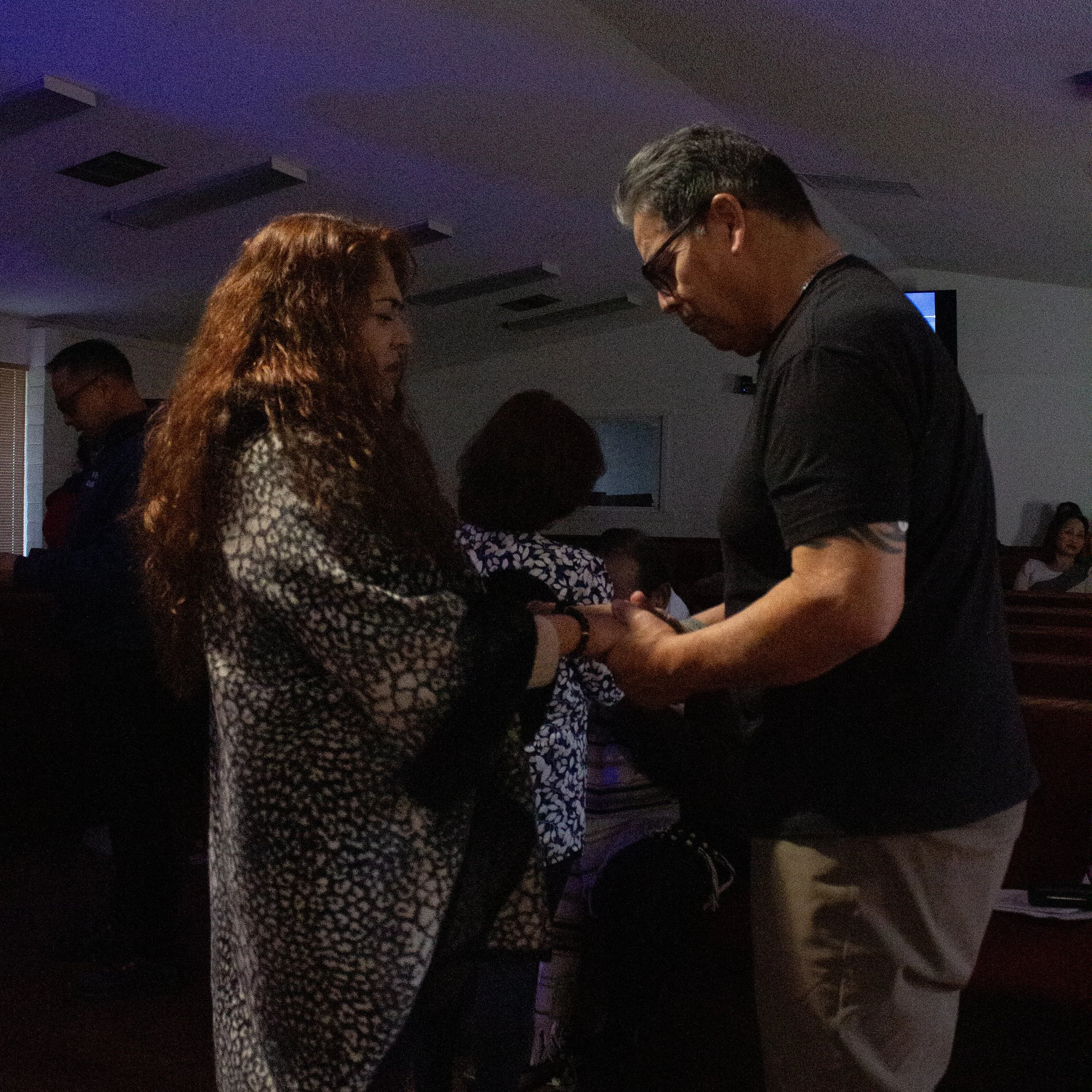 Two people, a man and a woman, are standing close together in a dimly lit room. The woman has curly hair and is wearing a patterned top, while the man has short hair, glasses, and is wearing a dark shirt. They appear to be engaged in a conversation or activity.