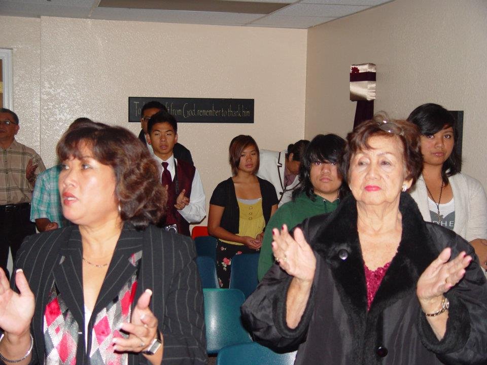 A group of diverse women and young people in a room, some clapping. The room has a blackboard with a quote, and the people are standing and appear to be engaged in an event or gathering.