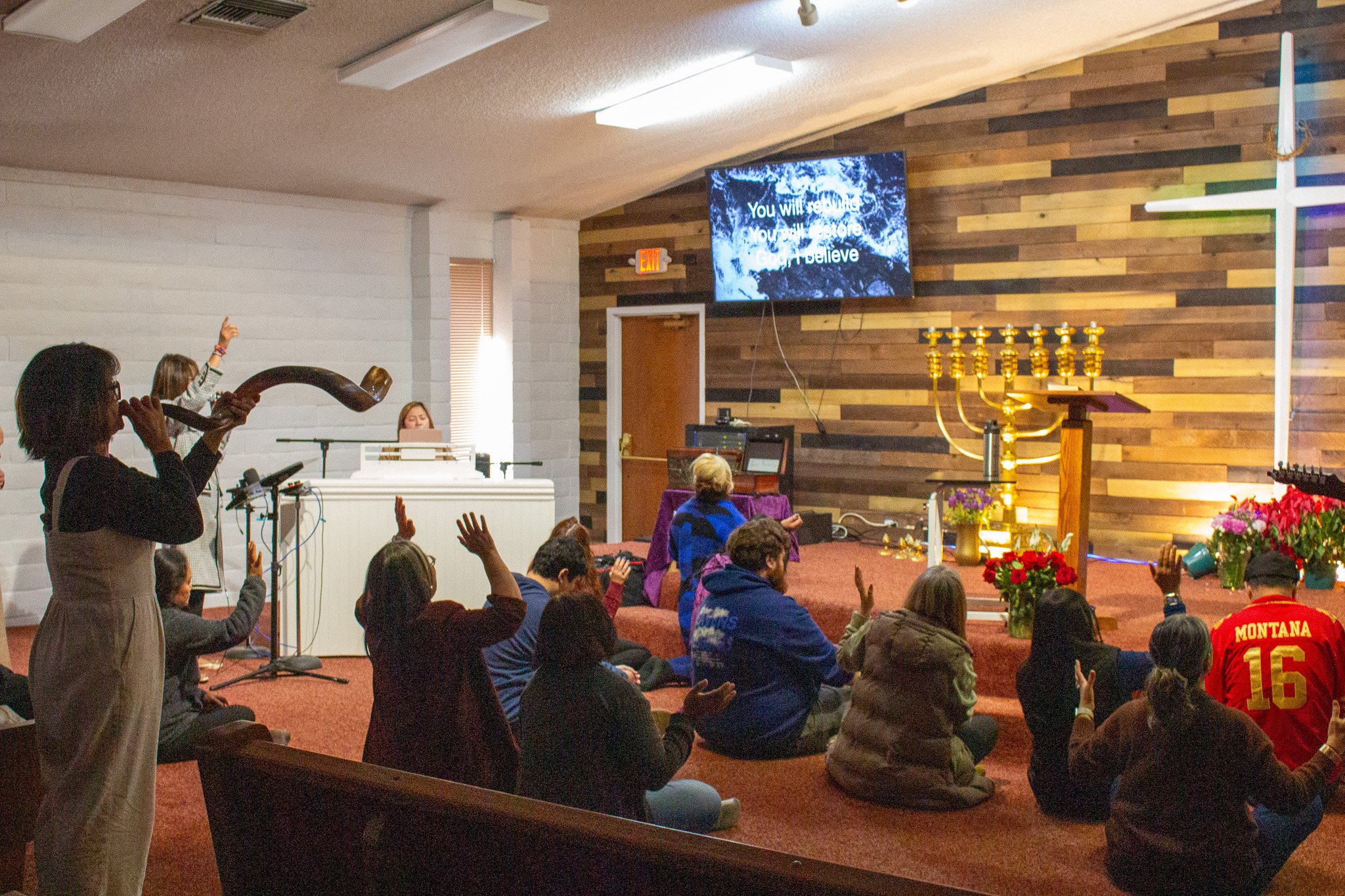 Church congregation seated on the floor during a service, with some people raising hands, a woman playing a horn instrument, and a large screen displaying a message, inside a church decorated with flowers and candles.