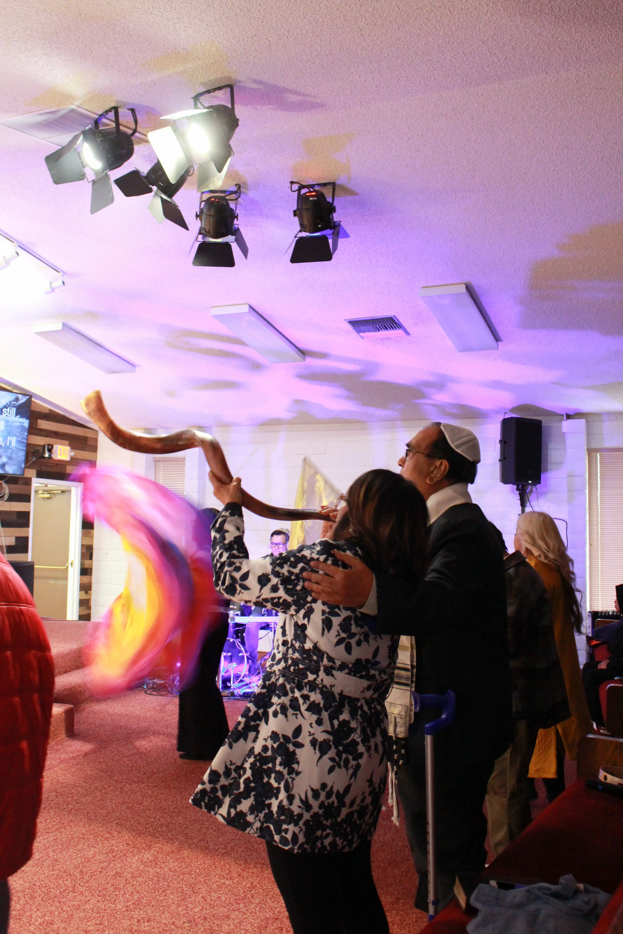 People dancing and celebrating at an indoor event. The scene shows a man and woman dancing closely, with the man wearing glasses and a dark jacket, and the woman in a black and white patterned dress. There are colorful decorations and stage lights overhead, creating a festive atmosphere.