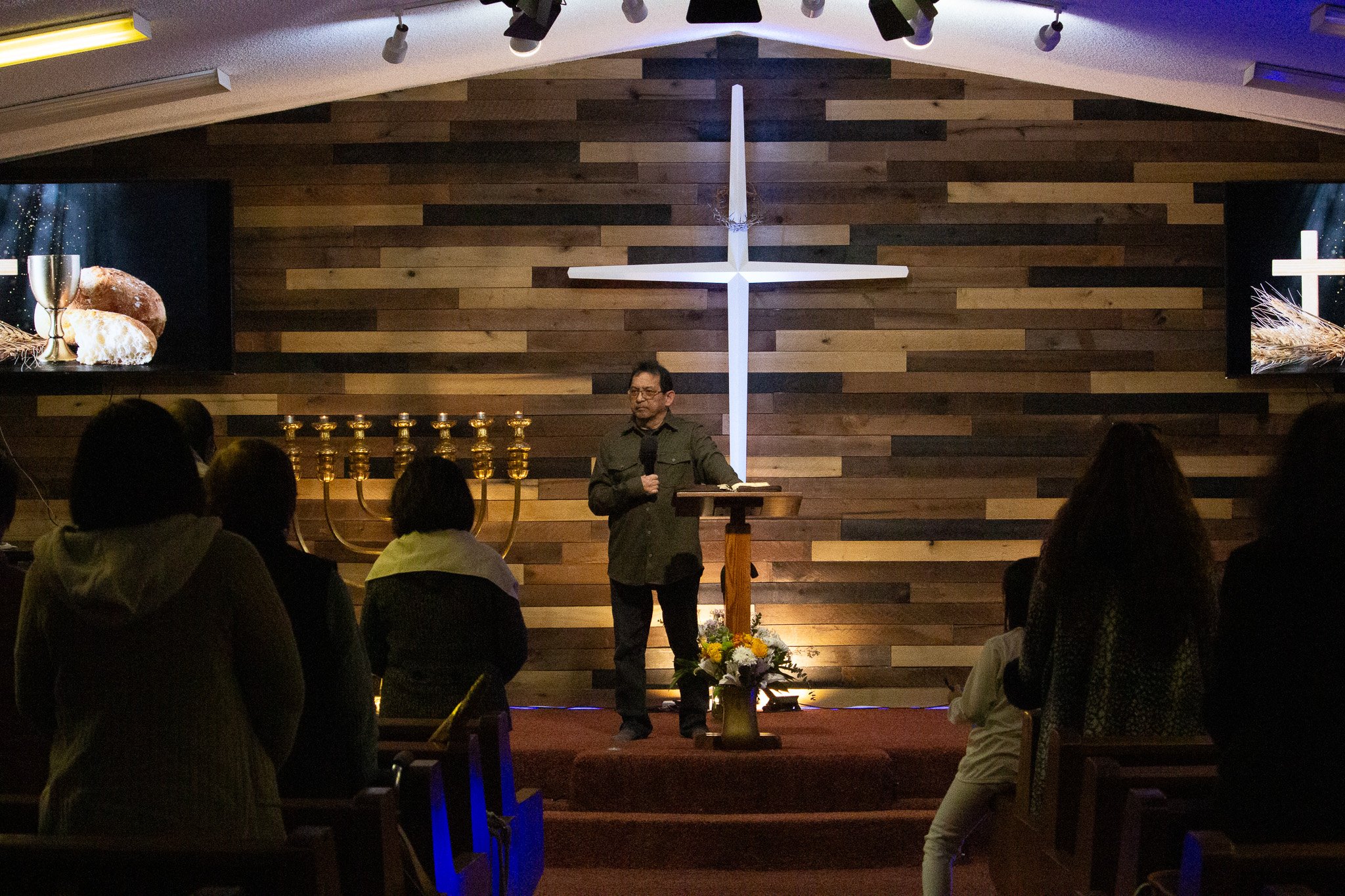A man speaks at a church pulpit in front of a large wooden cross during a church service. There are attendees seated in the audience, two screens display images of bread and wine to the church congregation.