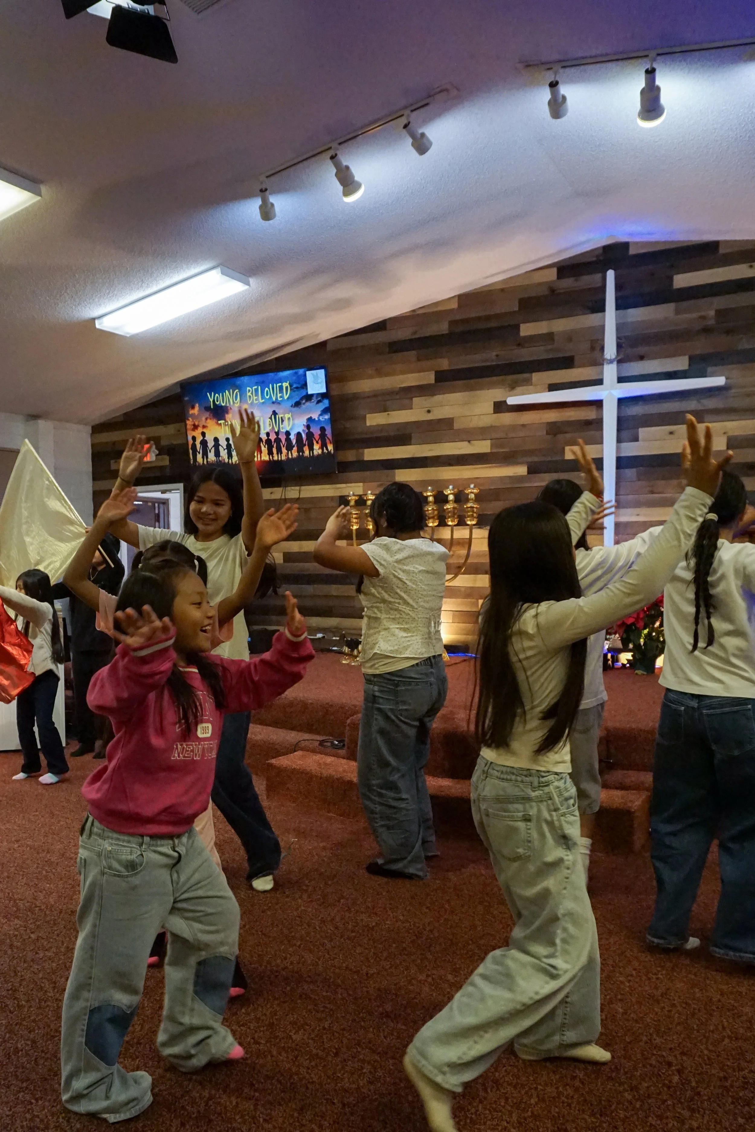 Children and adults dancing and celebrating inside a church. There is a large white cross on the wood-paneled wall, and a TV screen displaying the words 'YOUNG BELOVED' with silhouettes of children holding hands.