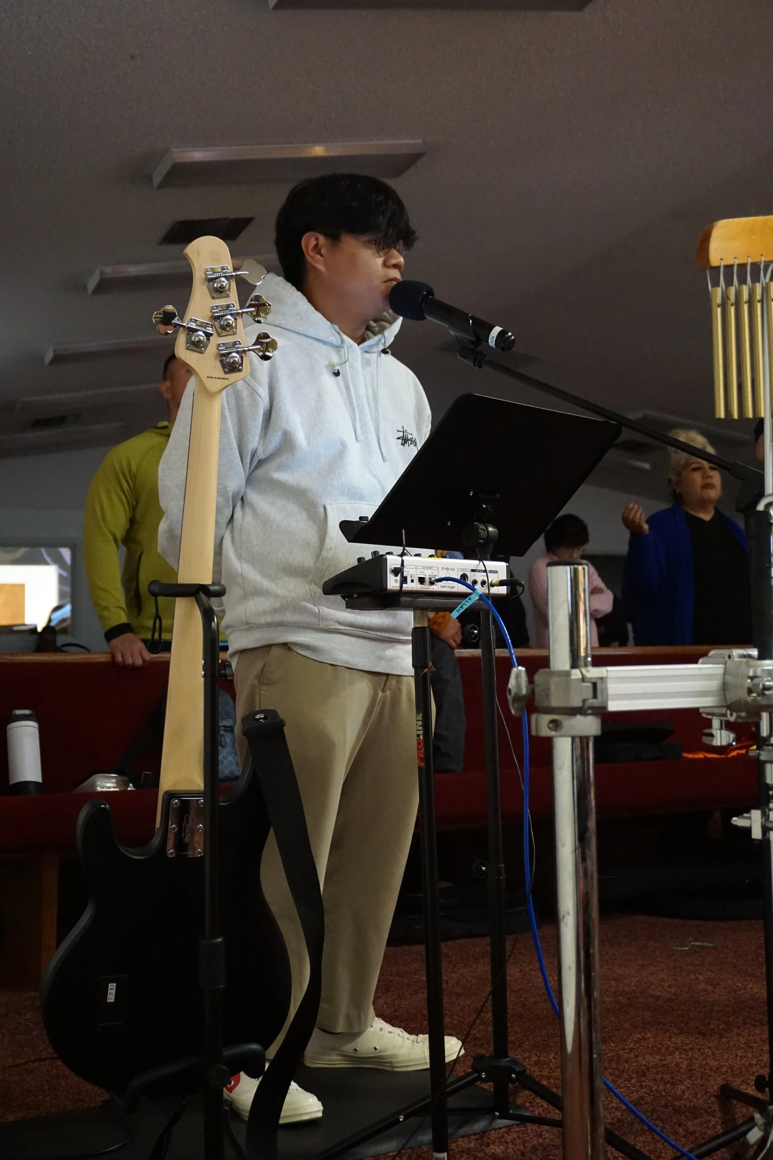 A young man with dark hair wearing a light gray hoodie and beige pants is standing on a treadmill, playing electronic music with equipment in front of him. He is speaking into a microphone. In the background, there are several people, some of whom are looking in his direction.