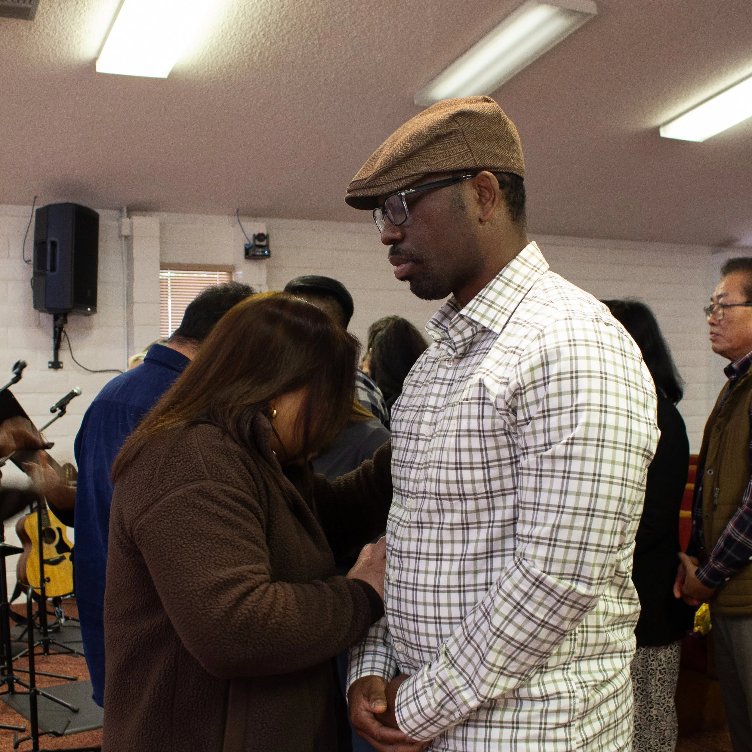 A man wearing glasses and a brown checkered shirt, standing with his head bowed, being prayed for by a woman in a brown sweater during a gathering in a room with musical instruments and a speaker in the background.