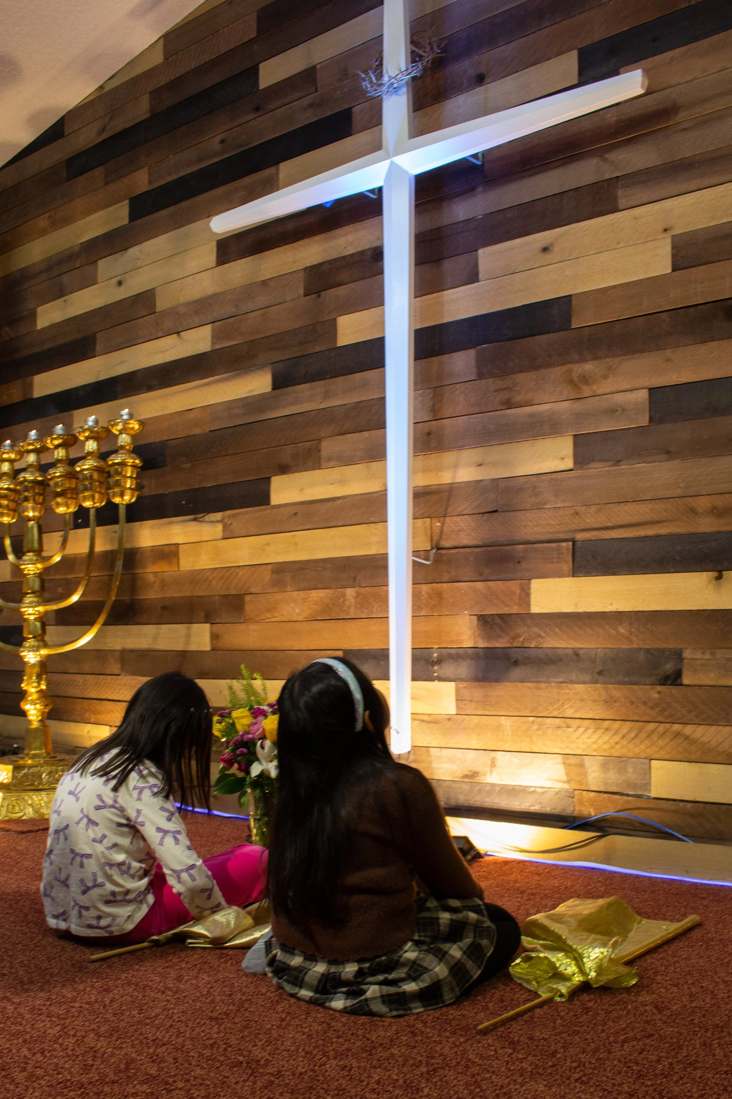 Children sitting on the floor during a religious or cultural ceremony, with a menorah, flowers, and a white candle in the background, in front of a wooden wall.