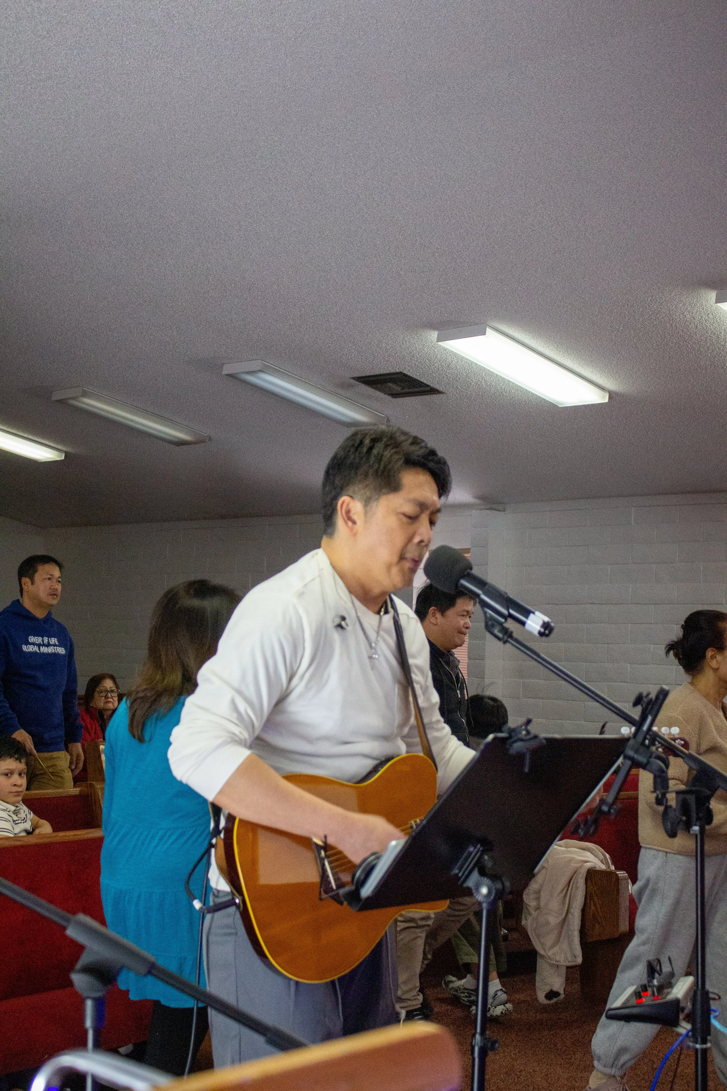 A man in a white shirt playing an acoustic guitar and singing into a microphone at an indoor event, with several people in the background, some sitting and some standing, including a woman in a blue jacket and a man in a blue hoodie.