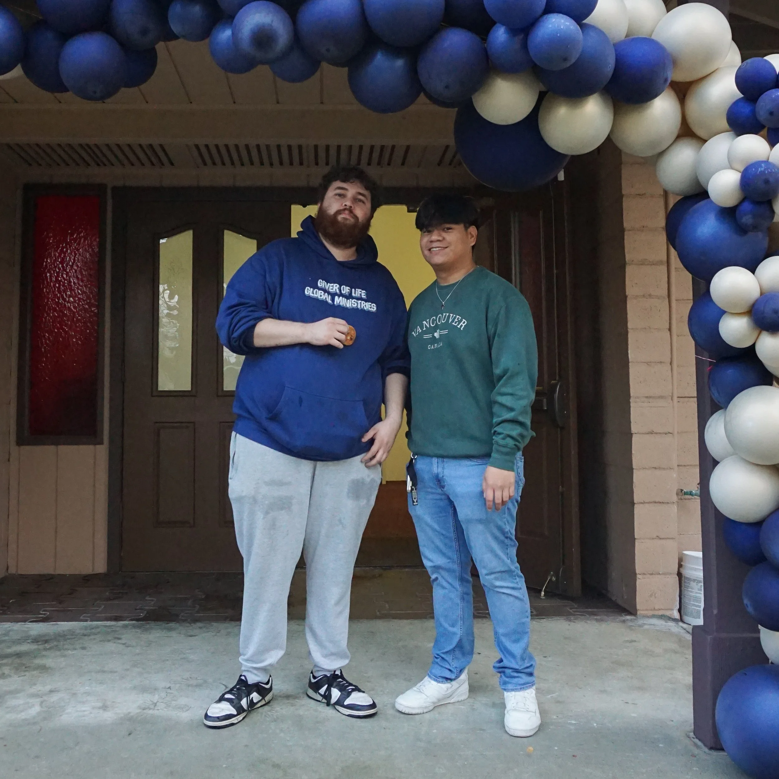 Two young men standing under a balloon arch, one wearing a blue hoodie and gray sweatpants, the other in a green shirt and blue jeans, both smiling.