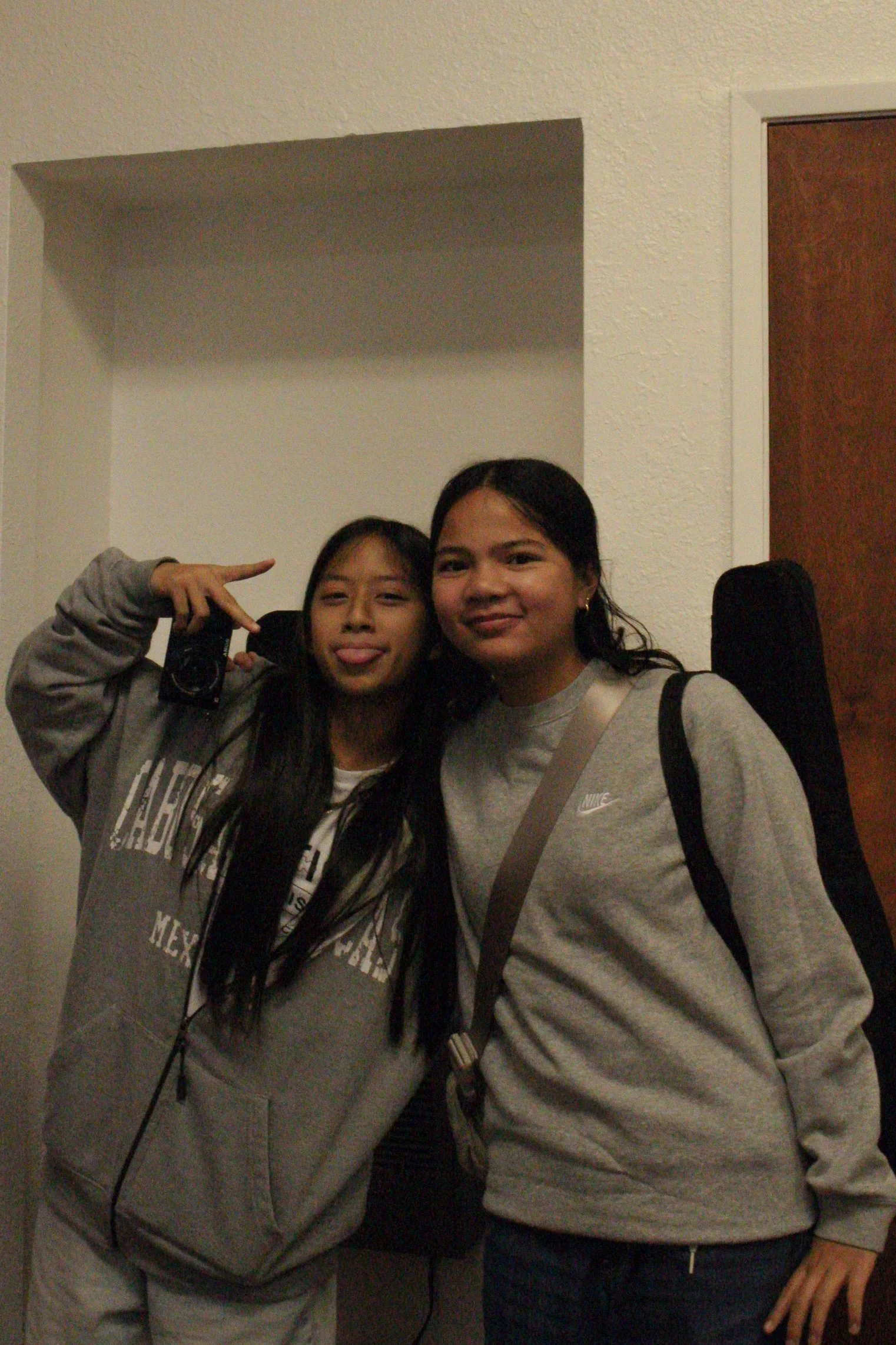 Two women taking a selfie in front of a wall. One woman is making a peace sign with her hand and sticking out her tongue. The other woman is smiling. They are both wearing casual clothing and have long dark hair.