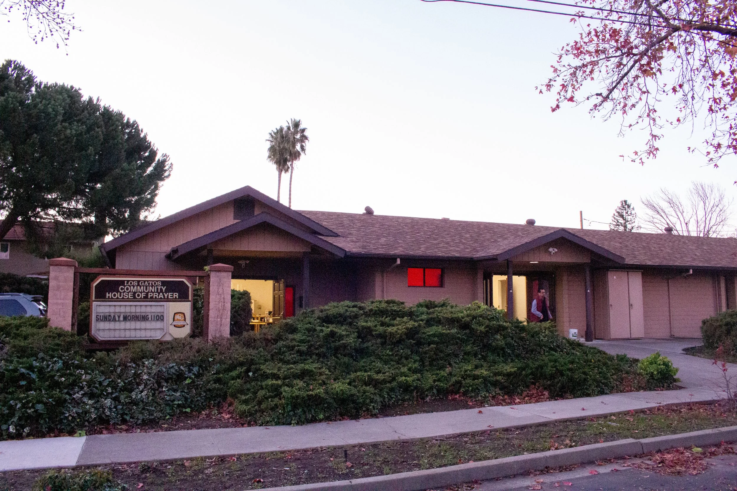 Church building with a sign in front reading "Los Gatos Community House of Prayer" and a smaller sign indicating Sunday morning services at 11:00. The sky is clear, and there are trees with fall foliage and palm trees around the building.