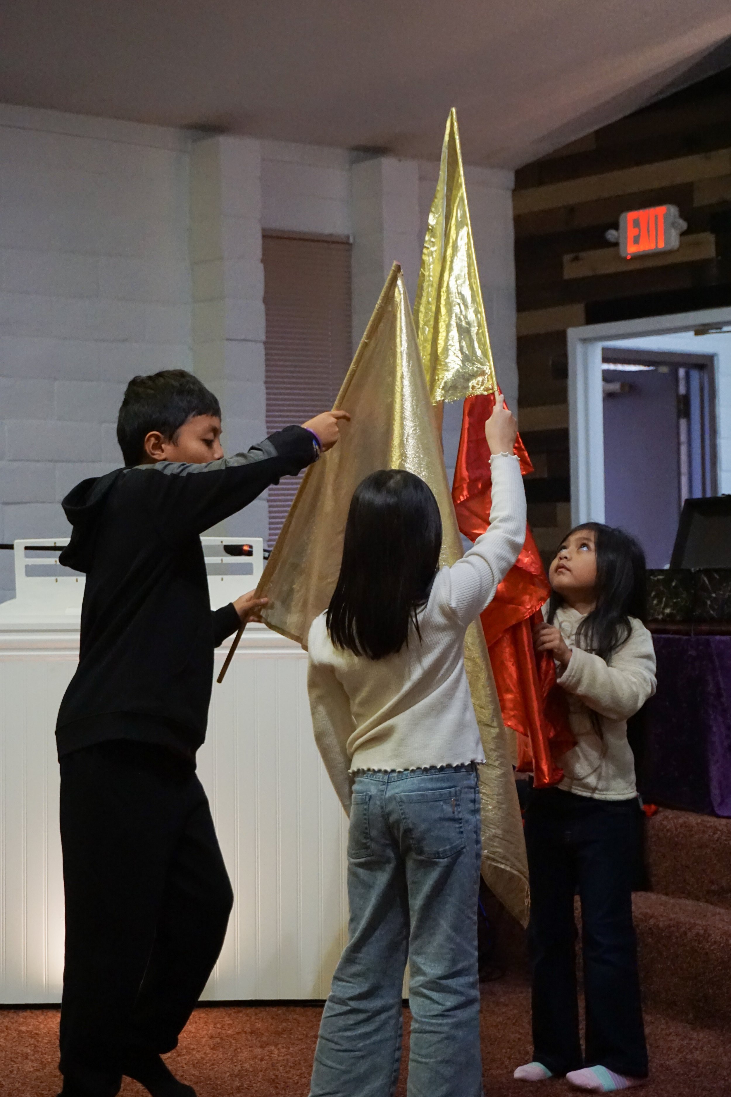 Children standing around a Christmas tree, some holding gold and red flags, in an indoor setting with brick and wood walls.