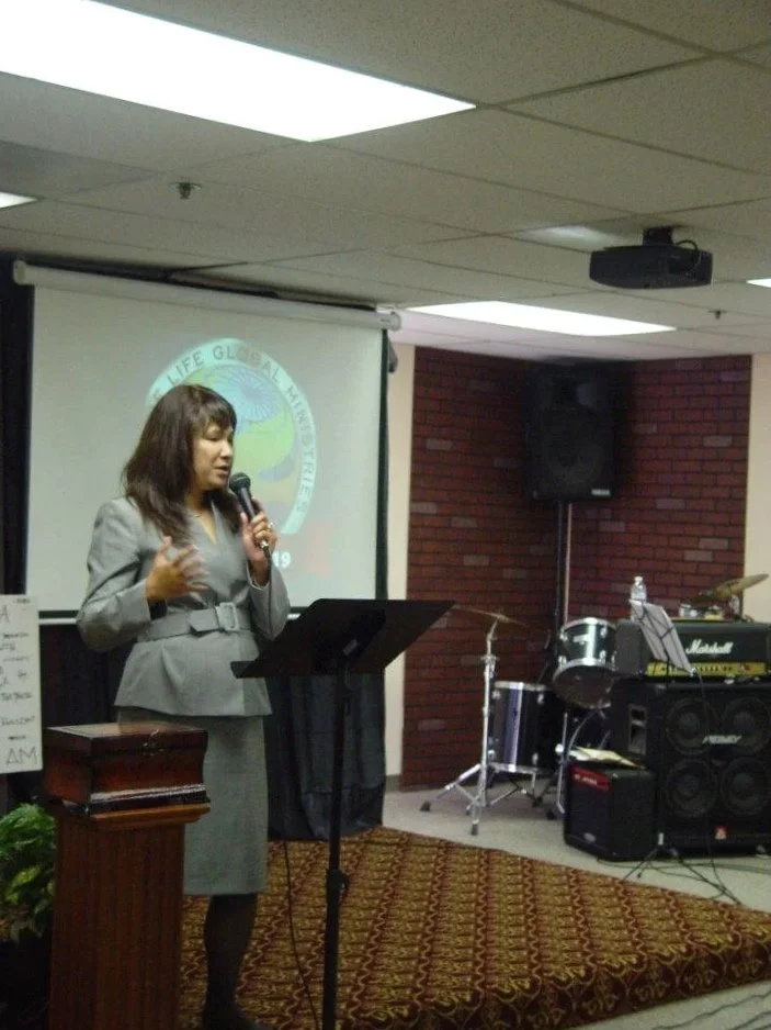 Woman in gray suit giving a presentation with a microphone in a conference room, featuring a projector screen, musical instruments, and a brick wall.
