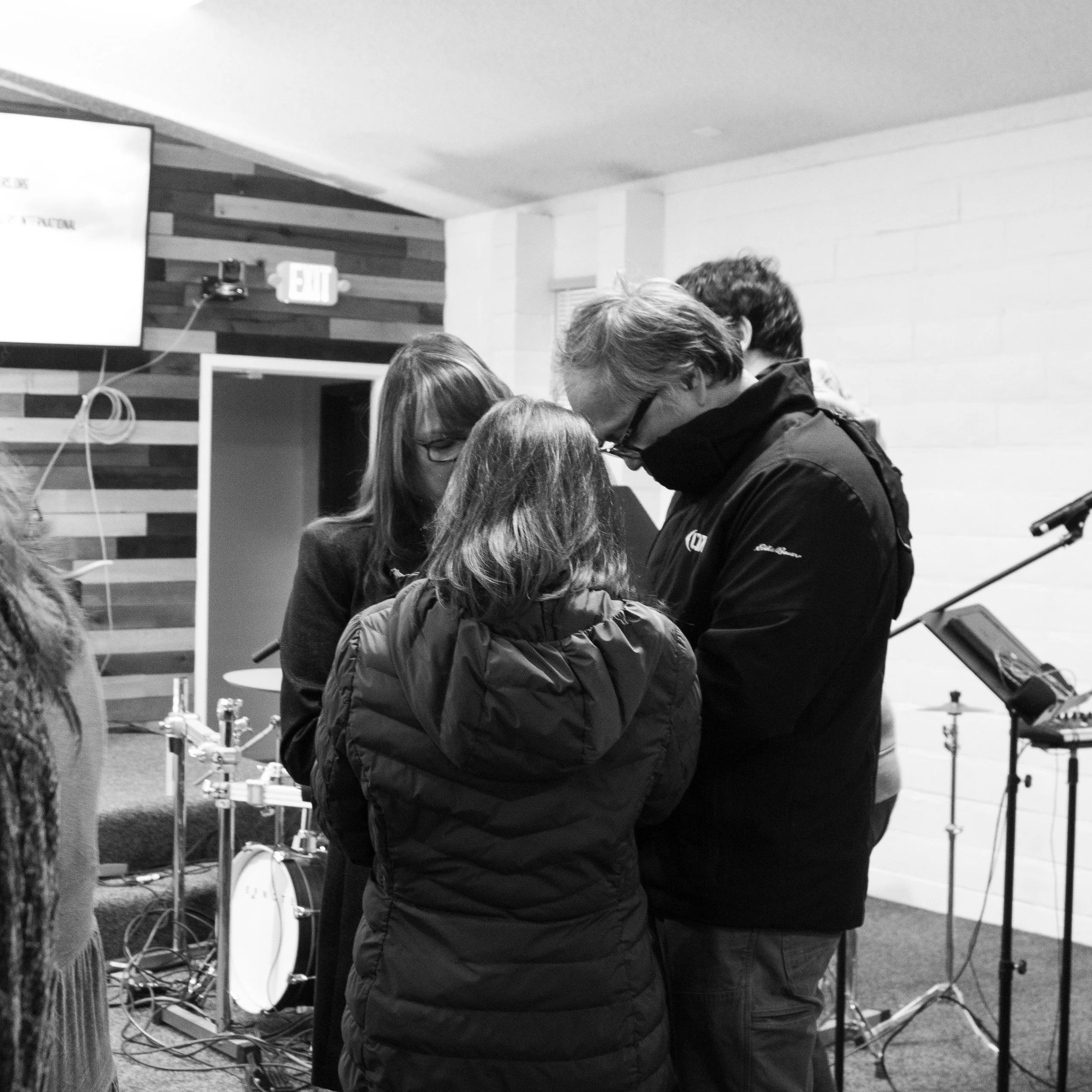 People gathered indoors, wearing winter clothing, with some looking at a screen or device, and a large television mounted on the wall in the background.