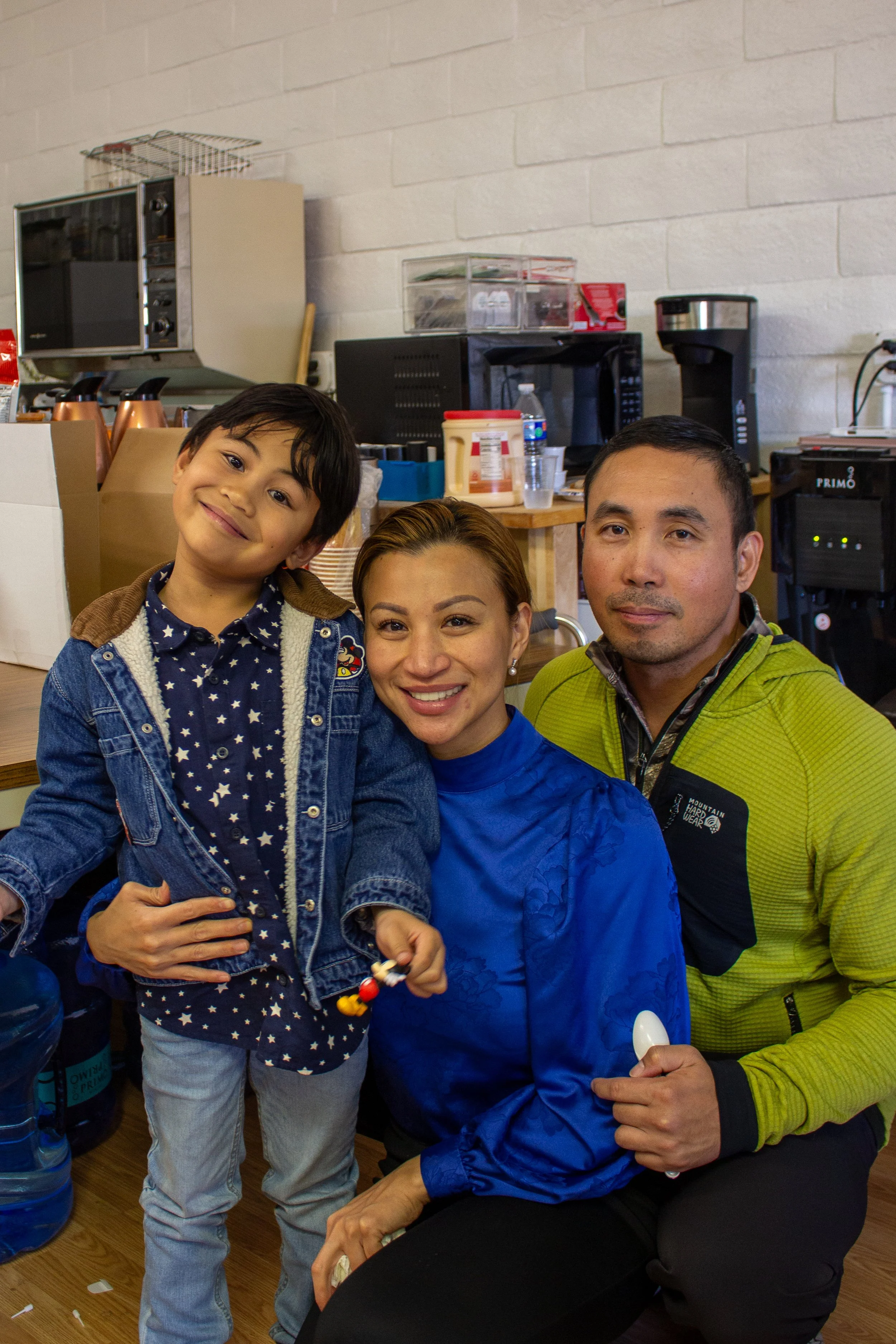 A young boy and a woman, with a man beside them, posing together in a kitchen or cafe setting, smiling at the camera.