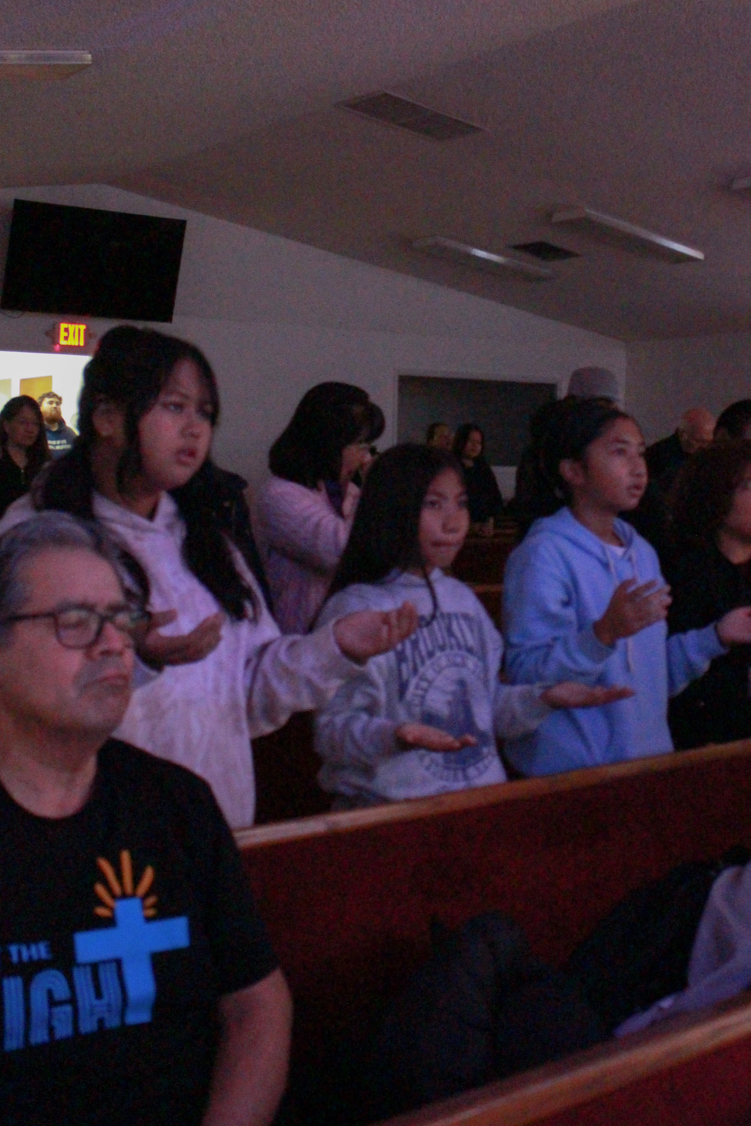 People standing in a church or religious setting, with their hands raised in prayer or worship. Some are wearing casual clothing, including hoodies and t-shirts.