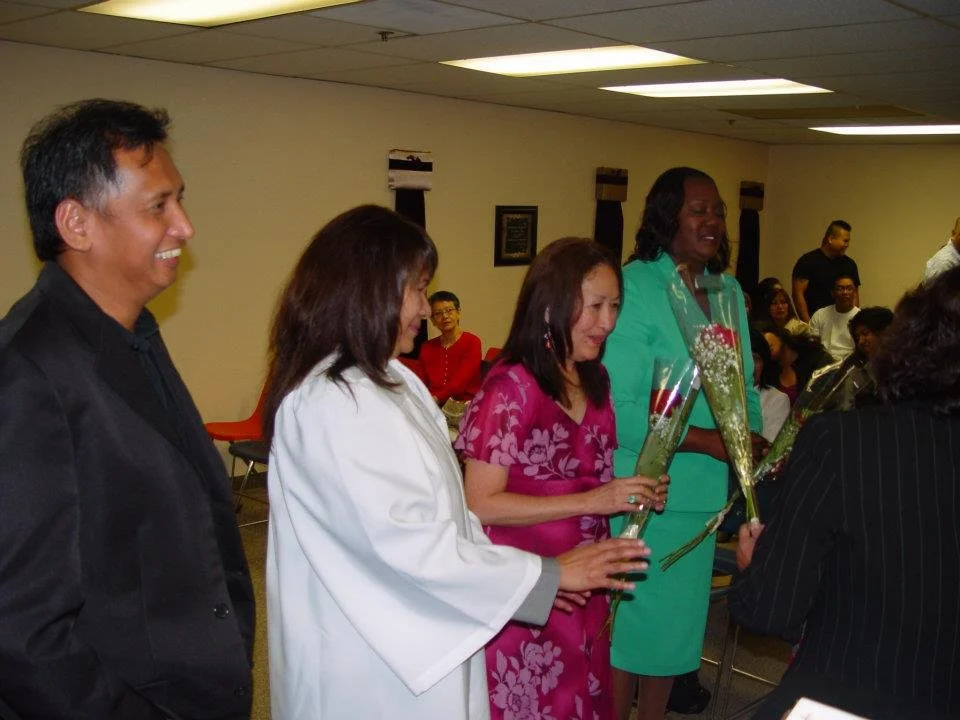 Four women and one man standing in a line inside a room, holding flowers, in a celebratory or formal event.