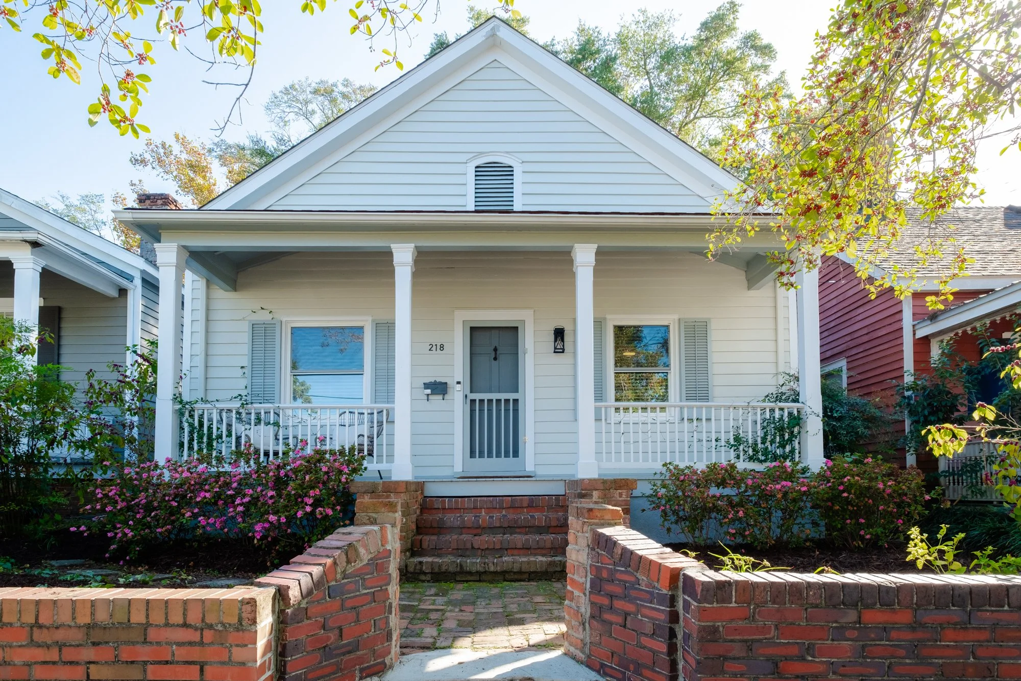 Front view of a white house with porch, covered stairs, and brick fence. Surrounding greenery and flowering bushes in front.