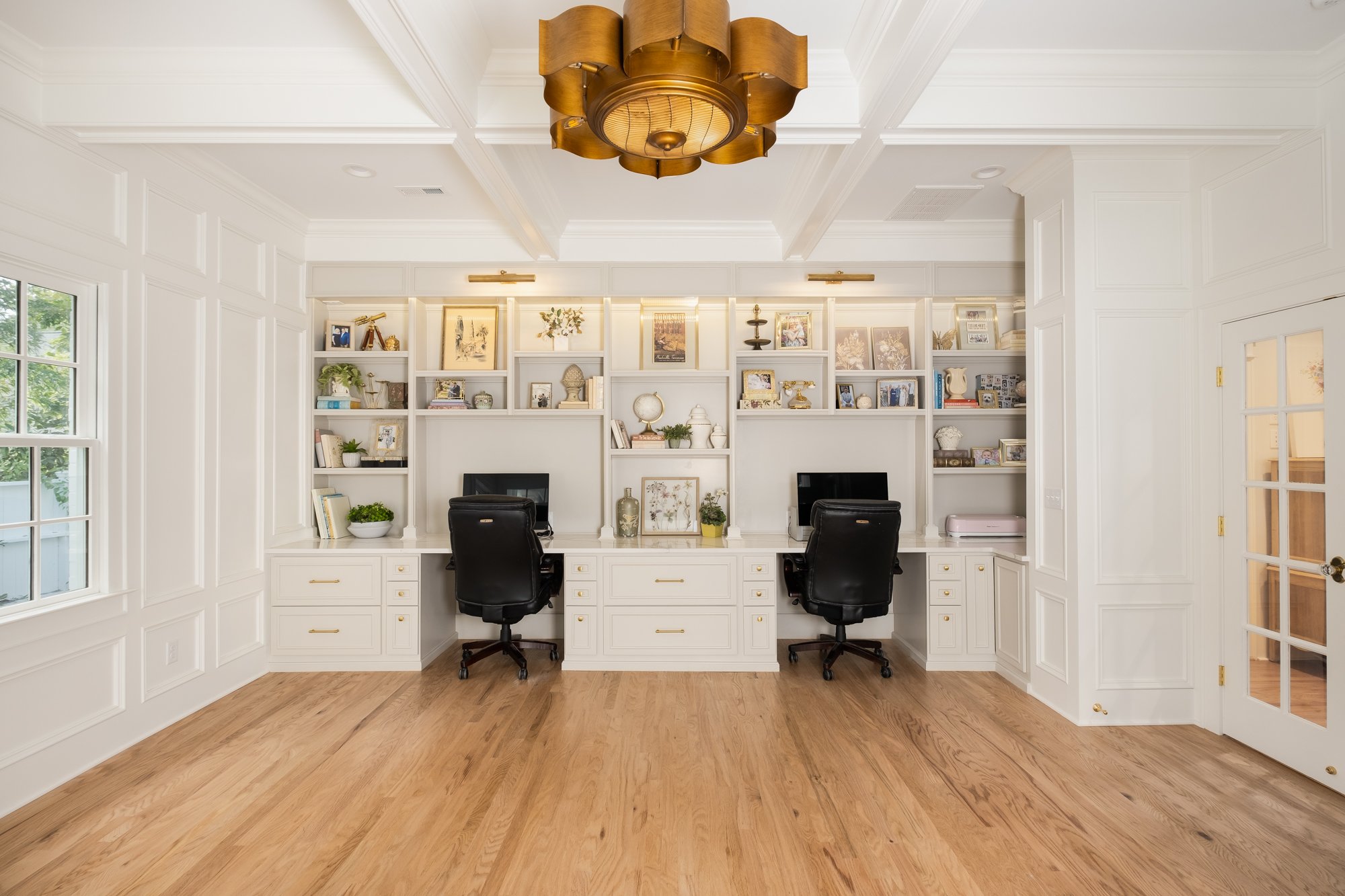 Home office with built-in white cabinetry, multiple shelves filled with books, framed photos, decorative objects, and two black office chairs. Room has white walls with wall paneling, hardwood floors, and a ceiling with a central bronze-colored chandelier.
