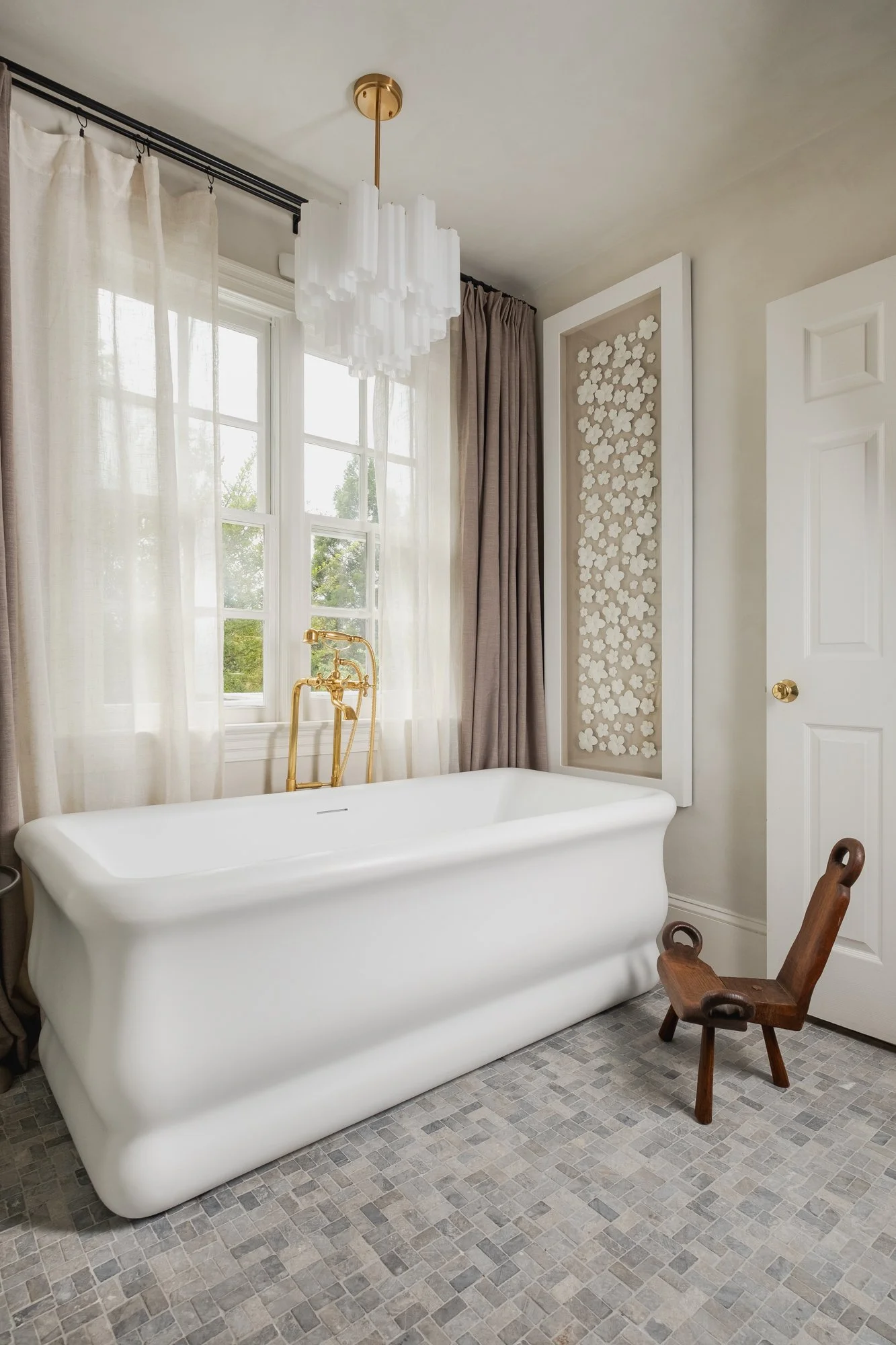 A bathroom with a traditional clawfoot bathtub, gold fixtures, and large windows with white and beige curtains. There's a wooden chair next to the tub and a decorative framed panel on the wall with white floral cutouts.
