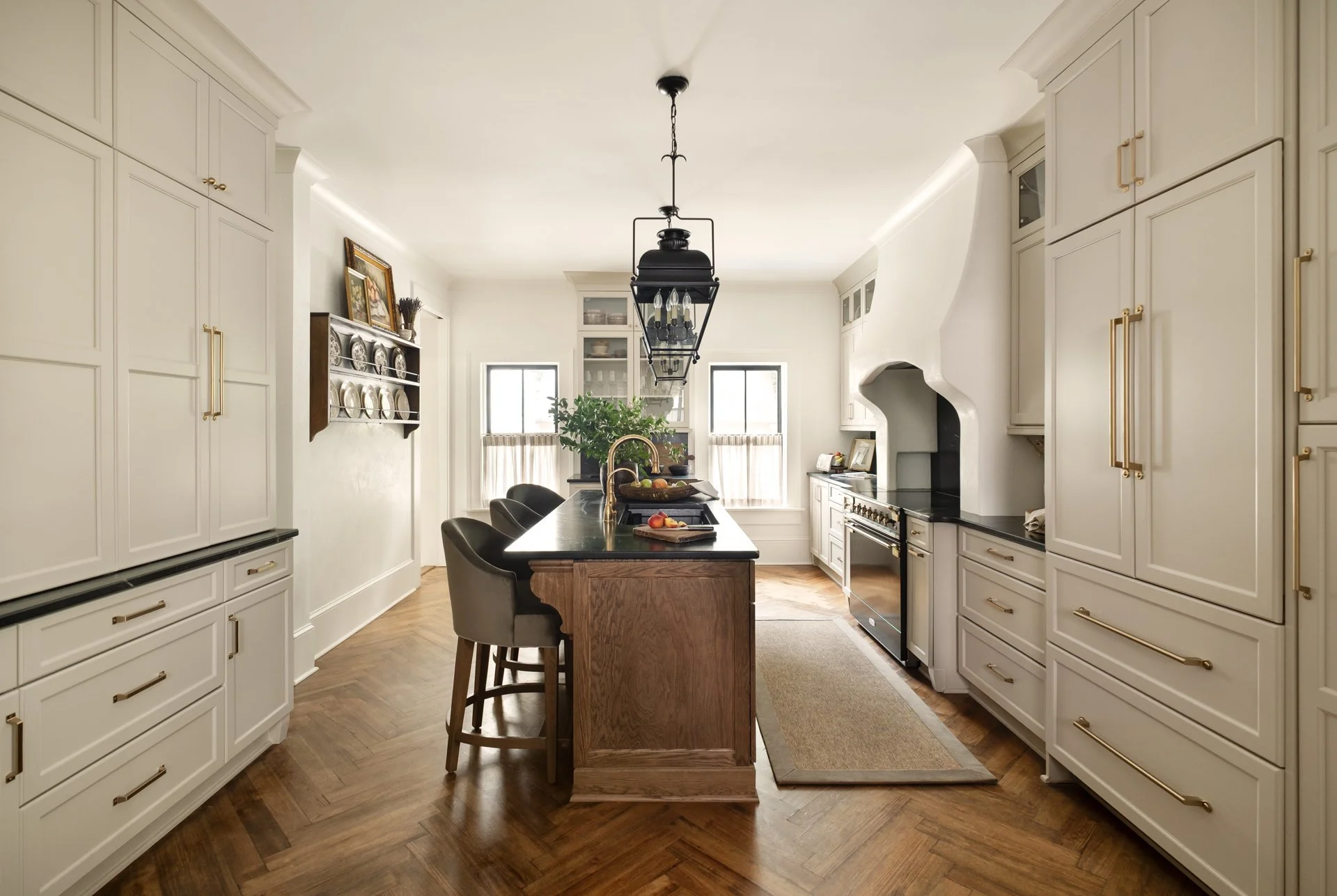 Bright kitchen with white cabinets, wooden island, black pendant light, and hardwood floors. Two windows with curtains, a plant, and decorative plates on a shelf add to the decor.