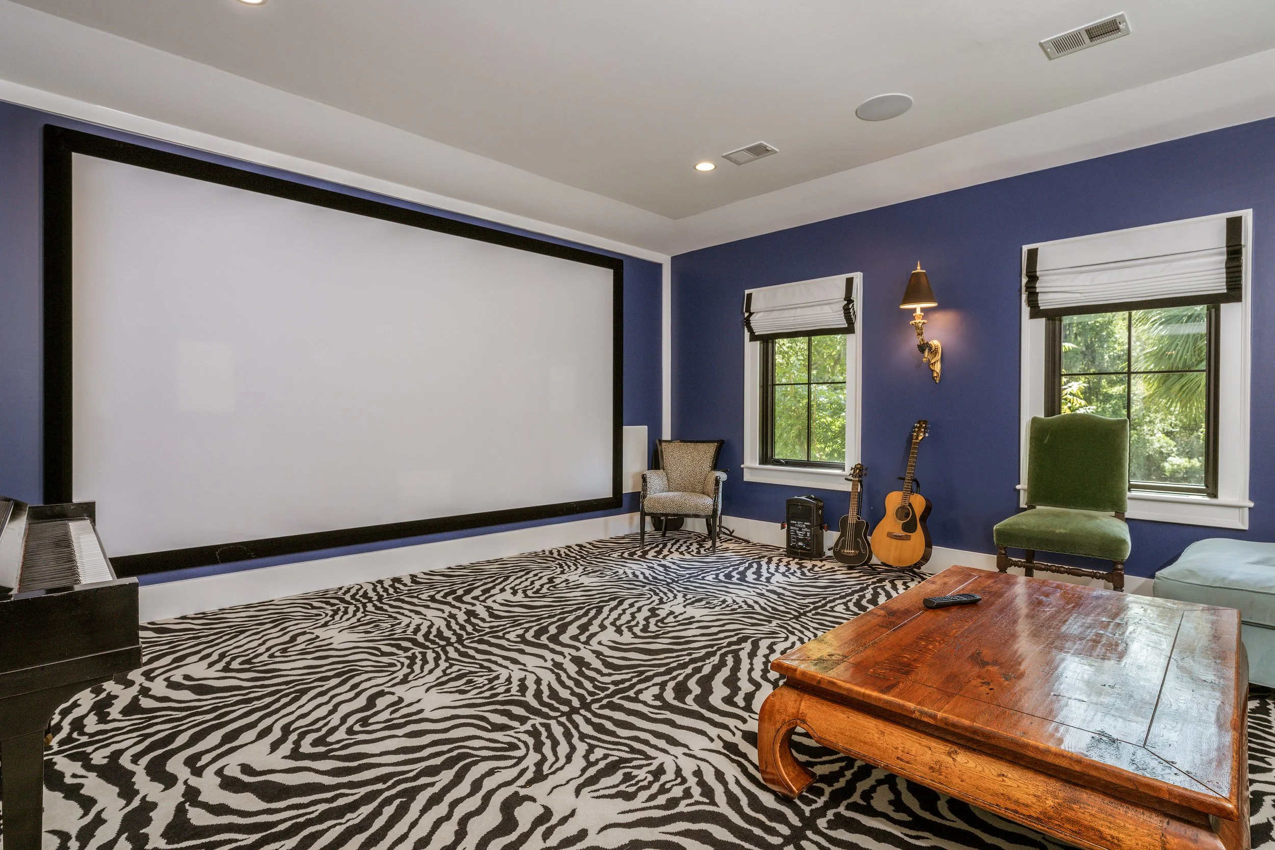 Luxury home media room with a large projection screen, blue walls, zebra-pattern carpet, built-in ceiling speakers, guitars, piano, and eclectic lounge seating.