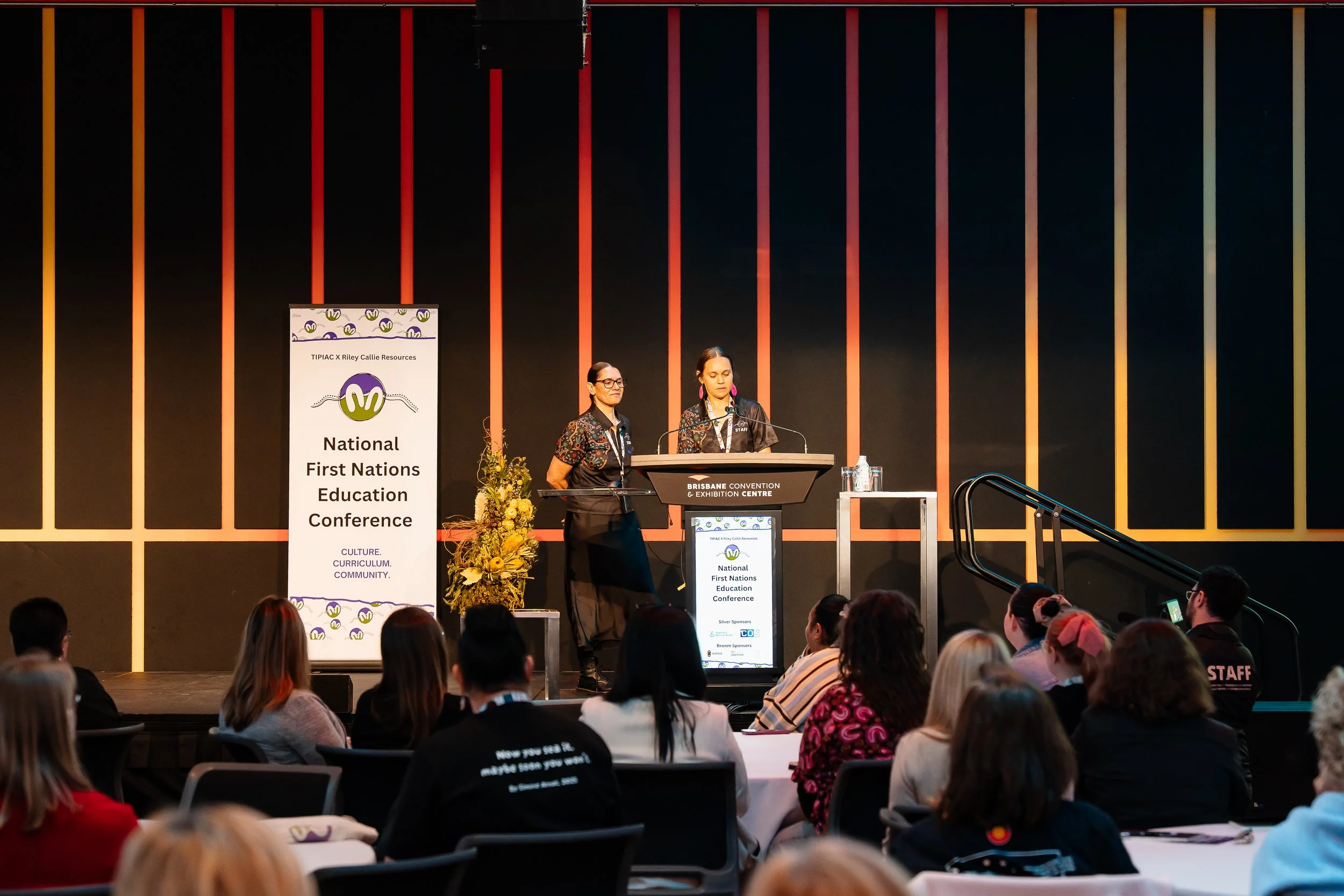 Women speaking at a conference stage with a banner that reads 'National First Nations Education Conference' and audience members sitting at tables in a large convention center.