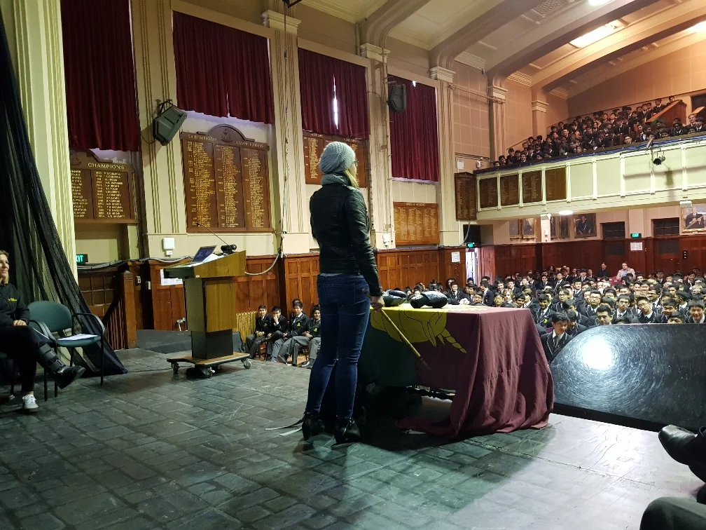 Vickie addresses a large audience of students in a school auditorium with wooden paneling and plaques on the walls.