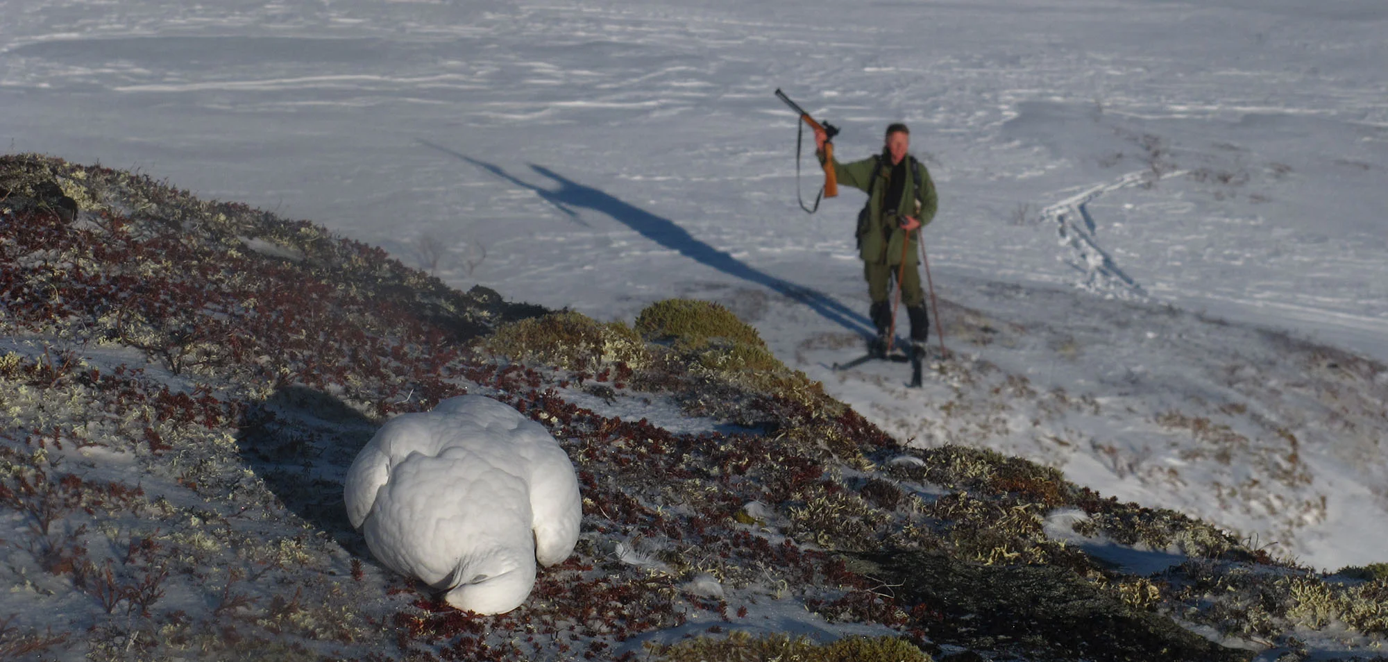 En person i fjellutstyr står i snøen og holder en jaktrifle opp i luften, på en snødekt fjelltopp med vinterlandskap.