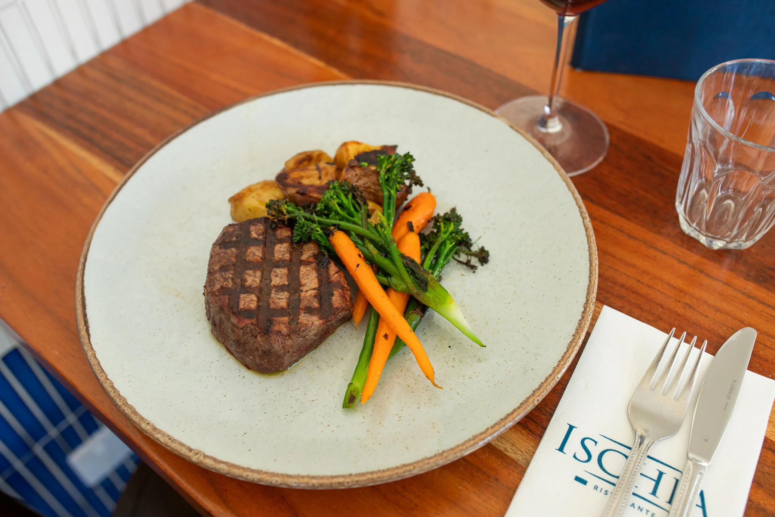 Plate with grilled steak, roasted potatoes, and sautéed carrots and broccoli on a wooden table, with a glass of red wine, glass of water, and silverware.
