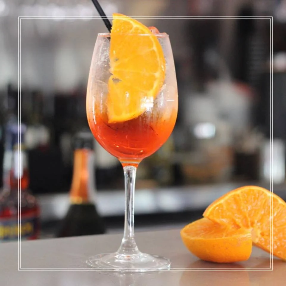 A cocktail glass with an orange wedge garnish and a straw, filled with an orange and red-colored drink, with additional orange wedges beside it.