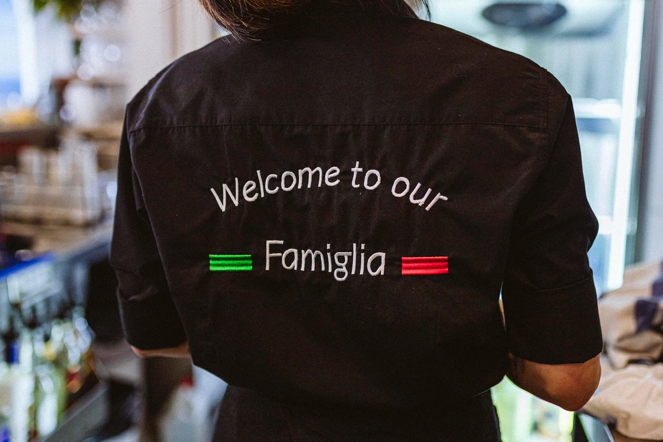 A person wearing a black jacket with white text and Italian flag colors that reads "Welcome to our Famiglia". The person is in a kitchen or cafe environment.