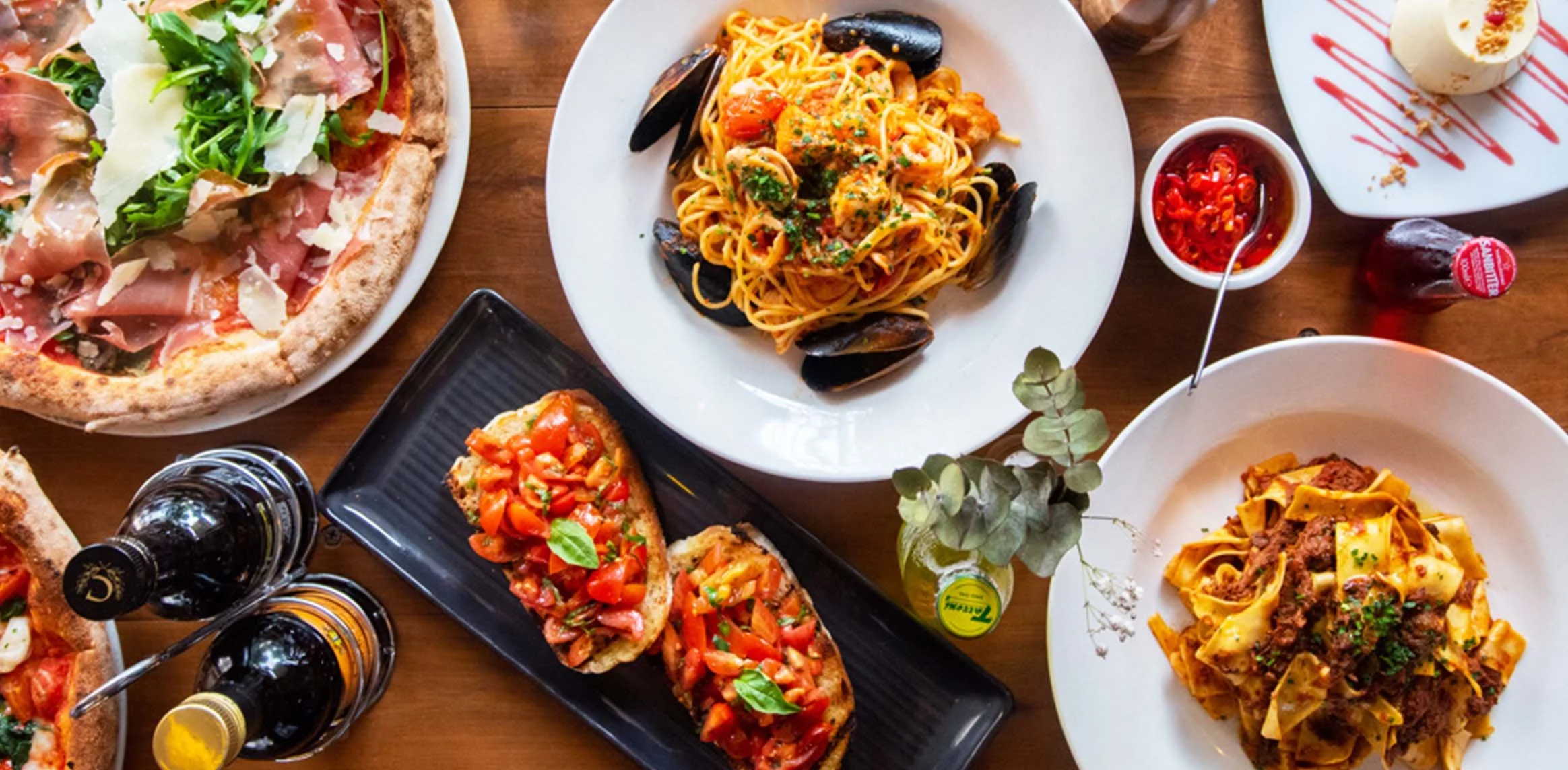 A variety of Italian dishes on a wooden table, including pizza with prosciutto and arugula, seafood pasta with mussels, bruschetta with tomatoes, and various sauces and condiments.