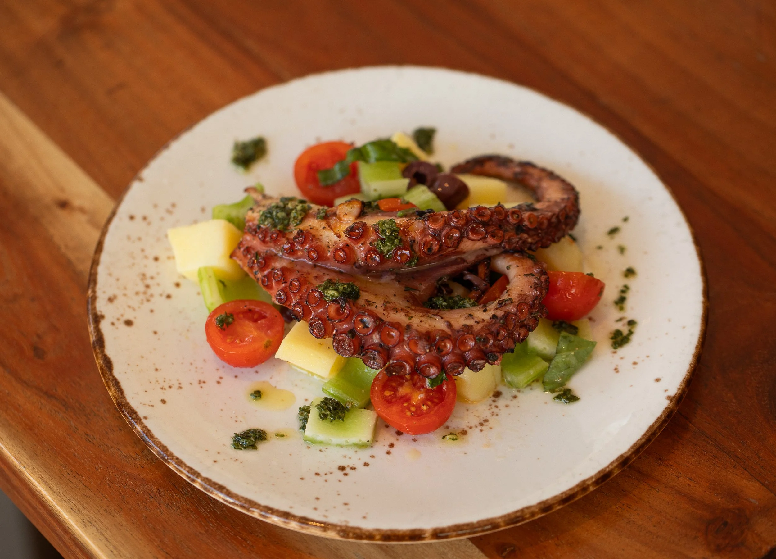 A plate of grilled octopus tentacle served with cherry tomatoes, potatoes, and herbs on a ceramic plate.