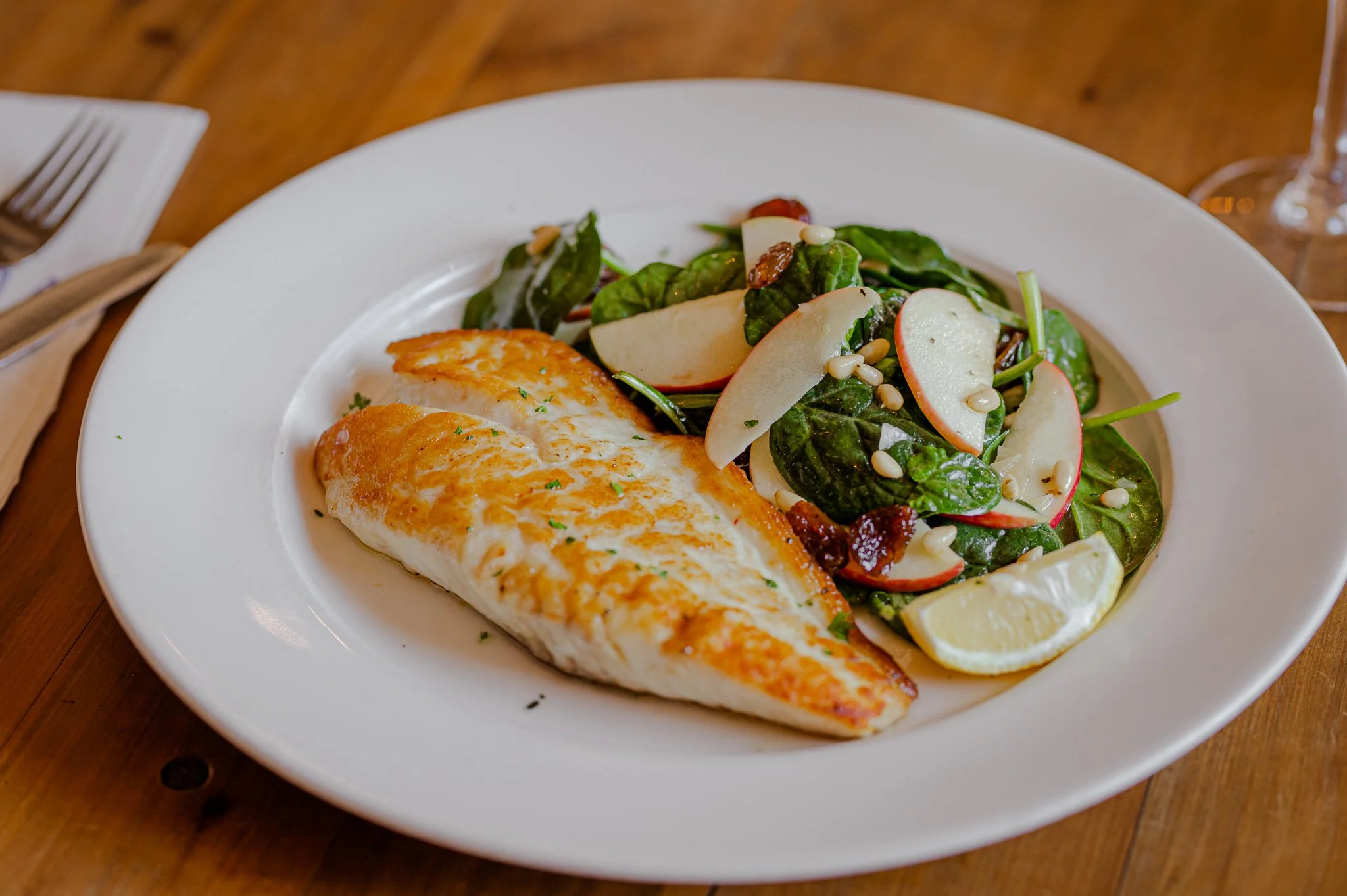 Plate of grilled fish with salad, apple slices, pine nuts, and lemon wedge on a white dish, on a wooden table.