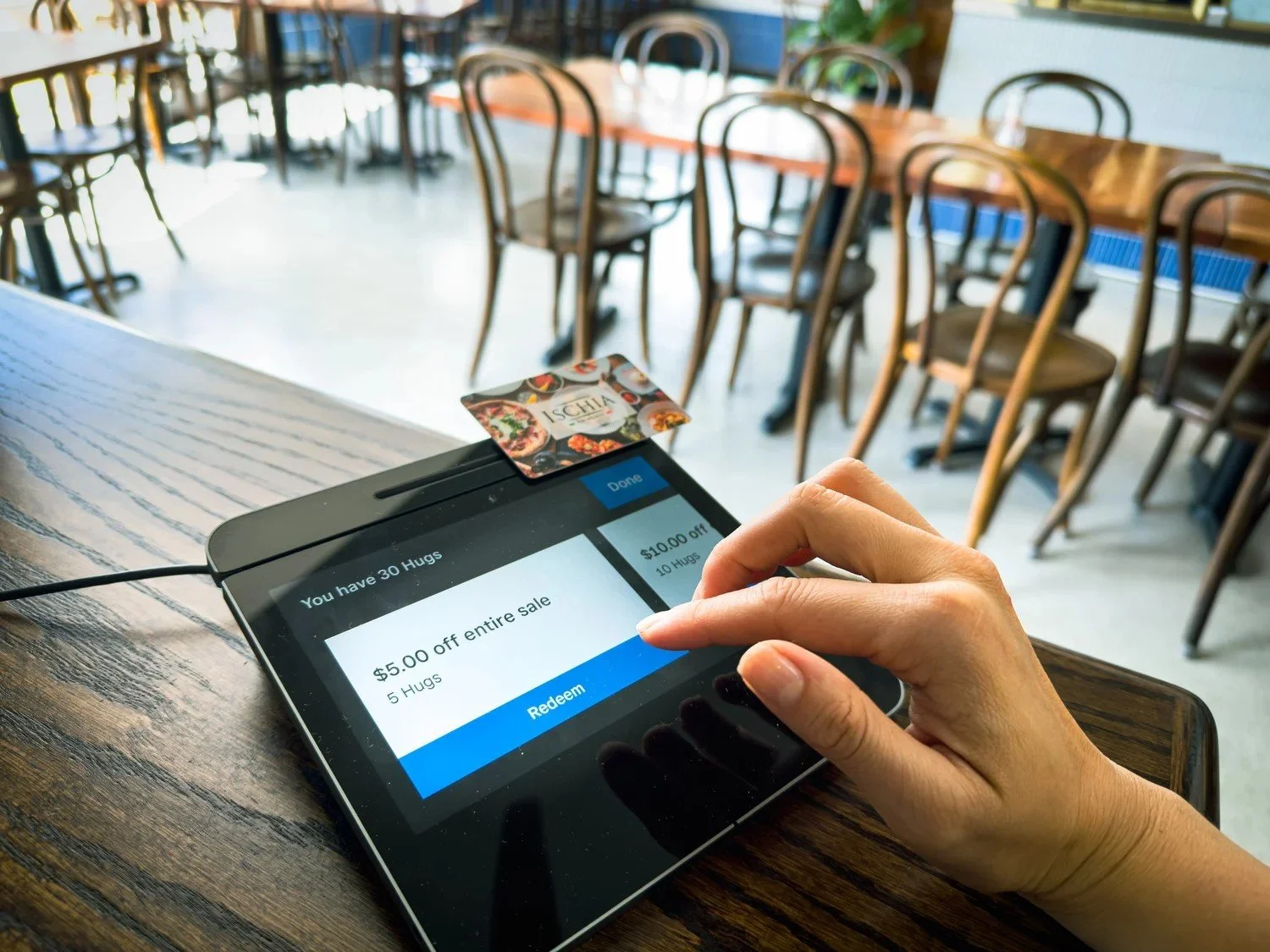 A person using a tablet on a wooden table, redeeming a coupon for $5 off an entire sale, in a restaurant or café with chairs and tables in the background.