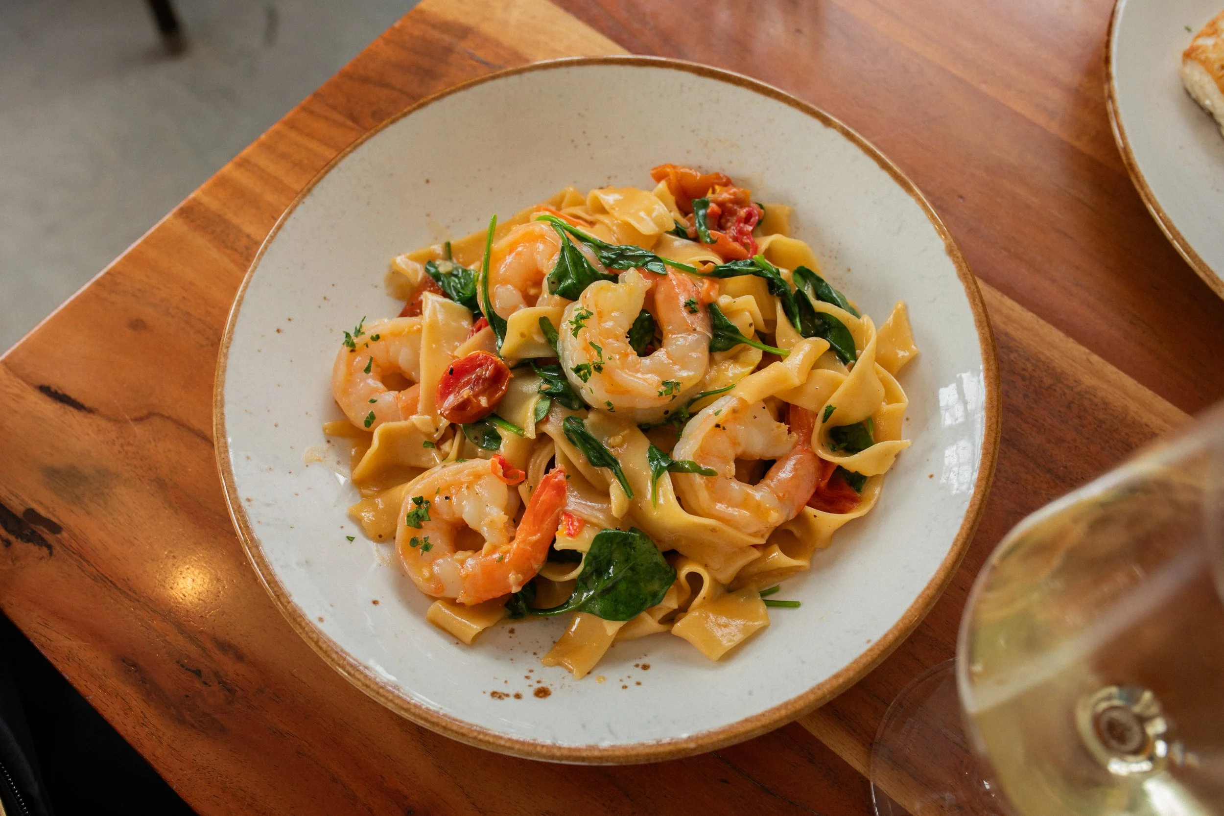 A plate of shrimp and pasta with spinach and cherry tomatoes on a wooden table, with a glass of white wine nearby.