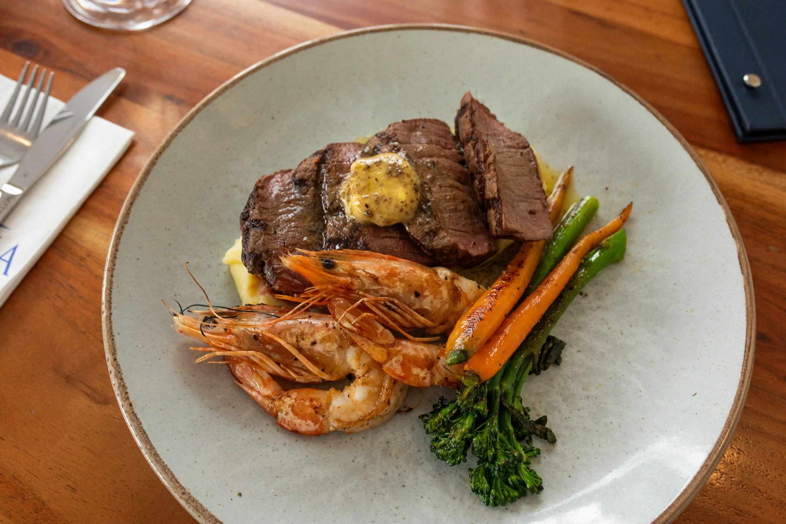 A plate with a cooked steak topped with a small dollop of butter, grilled shrimp, and a variety of cooked vegetables including carrots, broccoli, and green beans, on a wooden table.