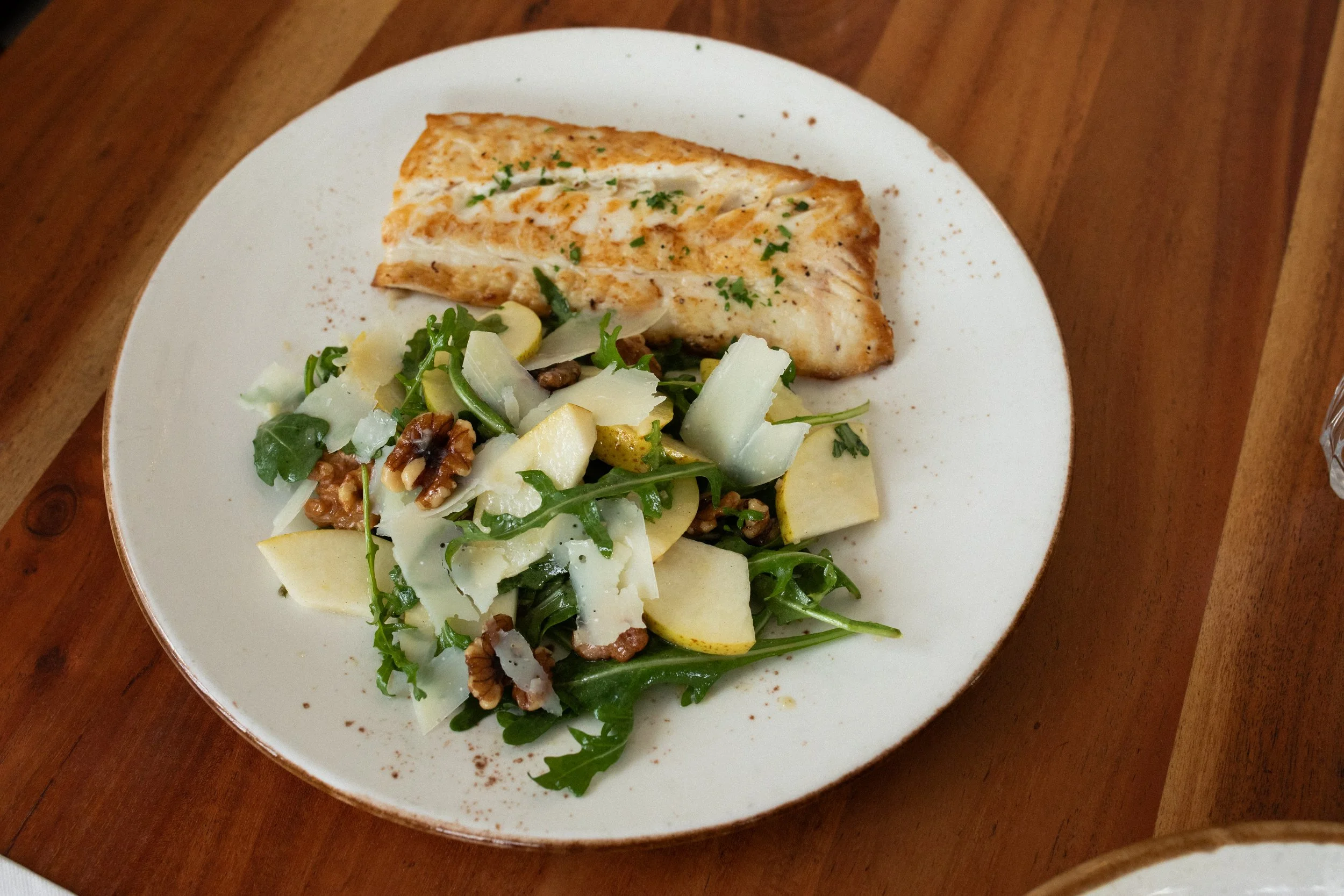 A white plate with a cooked fish fillet garnished with herbs and black pepper, served alongside a green salad with shaved cheese and chopped nuts, on a wooden table.