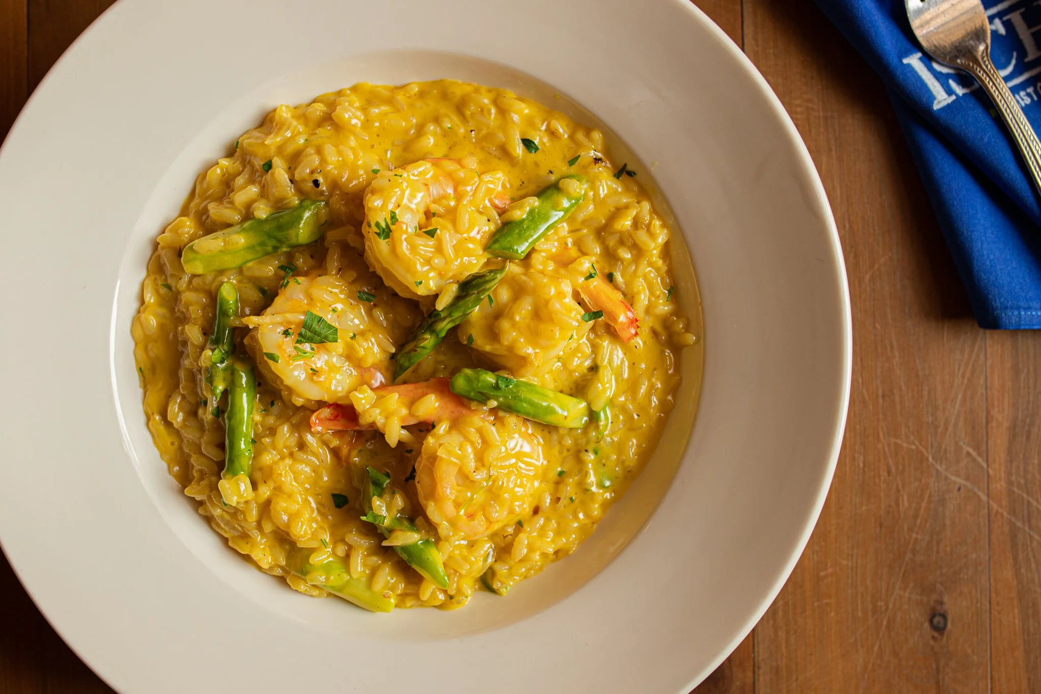A bowl of shrimp and asparagus risotto garnished with chopped parsley on a wooden table, with a blue napkin and silver spoon nearby.