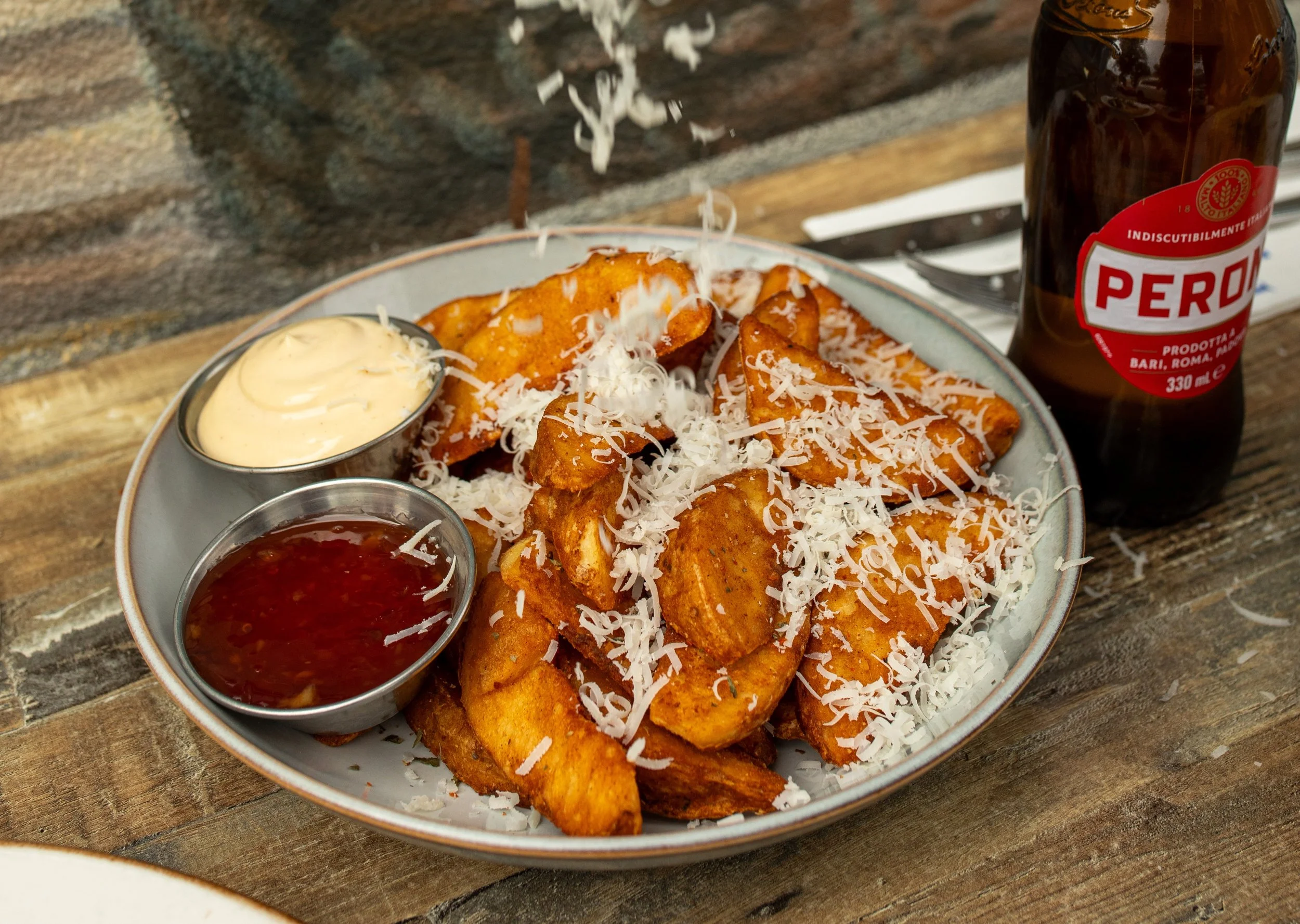 Plate of fried fish fillets topped with shredded cheese, served with small cups of tartar sauce, cocktail sauce, and salsa, placed on a wooden table next to a bottle of Peroni beer.