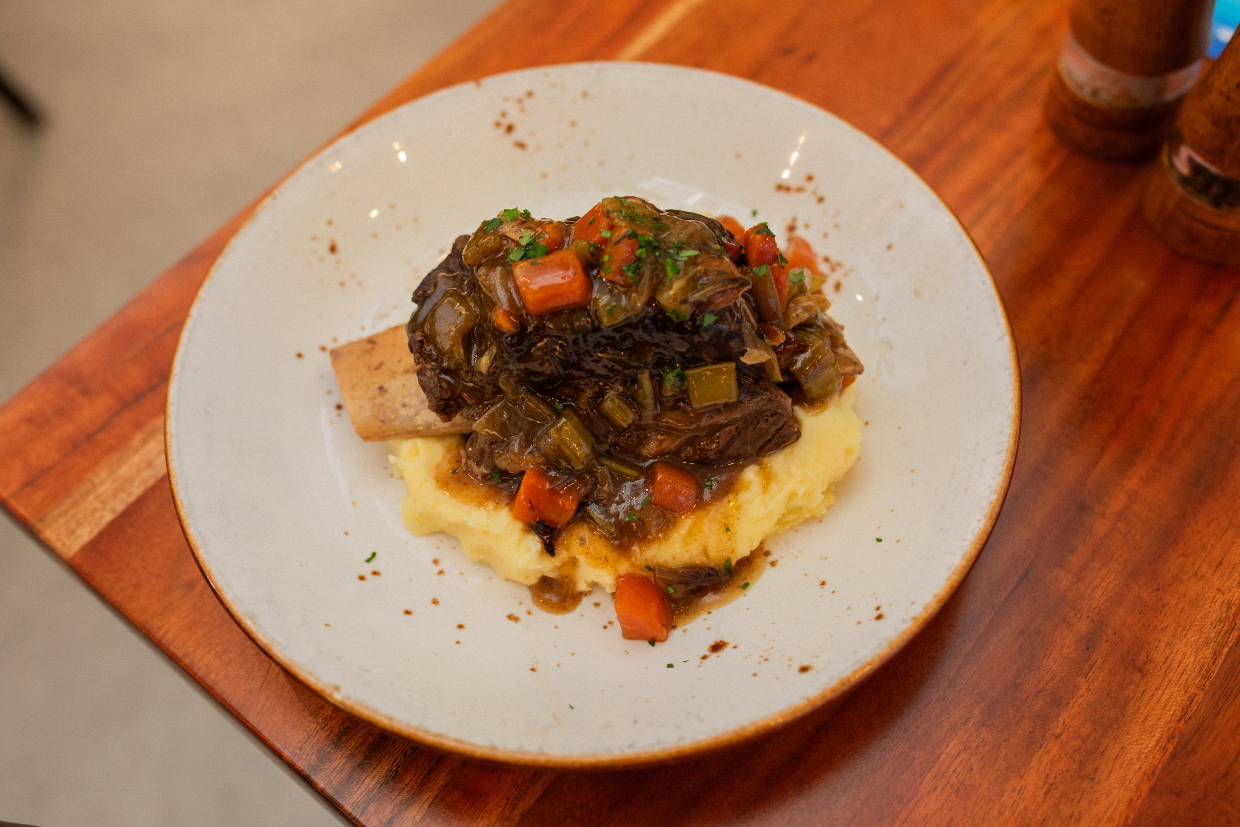 A plate of mashed potatoes topped with beef stew containing carrots, celery, and green herbs, on a wooden table.